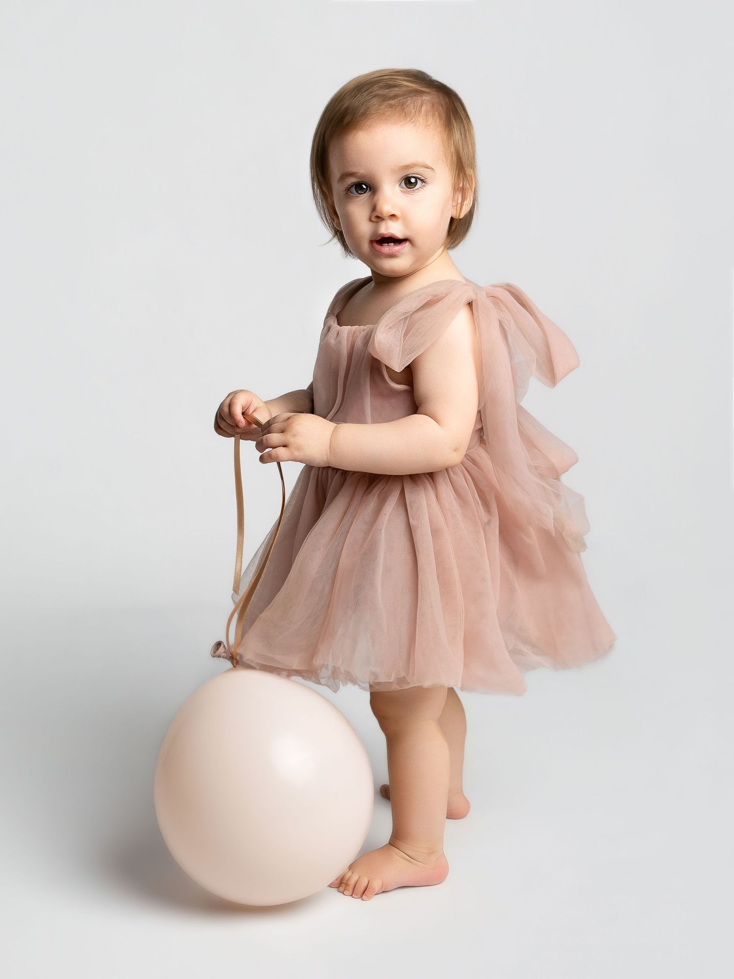Toddler girl in blush pink tulle dress holding a balloon string, standing barefoot against white background.