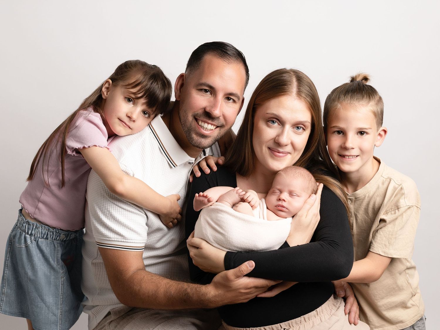 Happy family of five posing for a newborn portrait, parents holding baby with two young children smiling against white background.