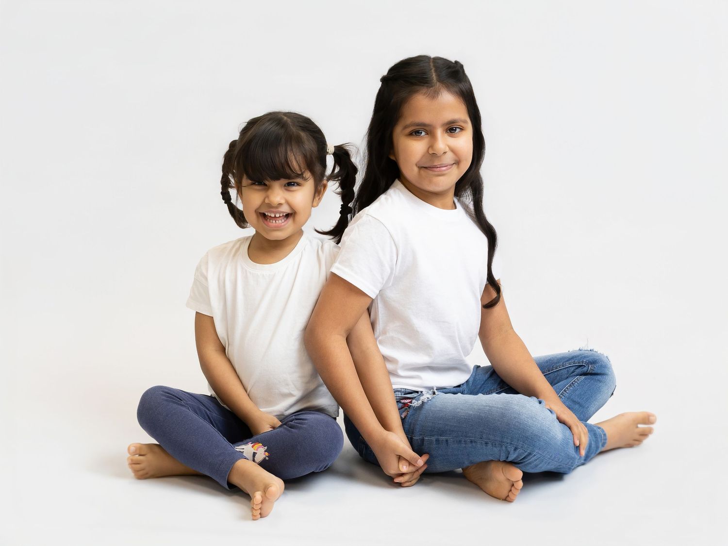 Two young girls in white t-shirts and jeans sitting cross-legged back to back, smiling at camera on white background.