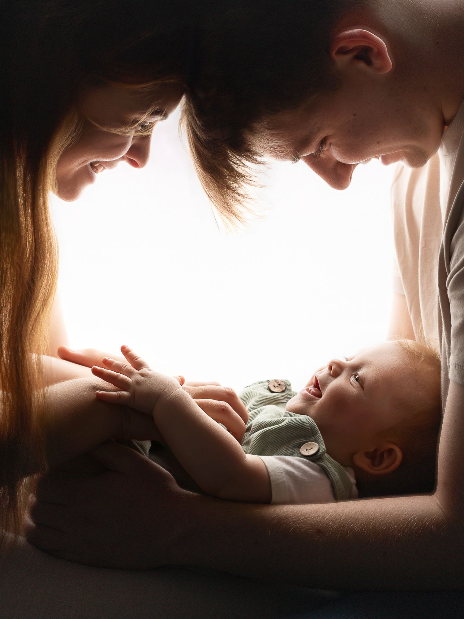 Two parents leaning over their smiling newborn baby, bathed in soft glowing backlight in an intimate family portrait.