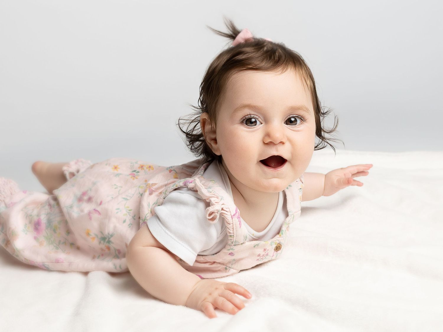 Adorable baby girl in floral romper lying on tummy, smiling brightly during a professional photo shoot.