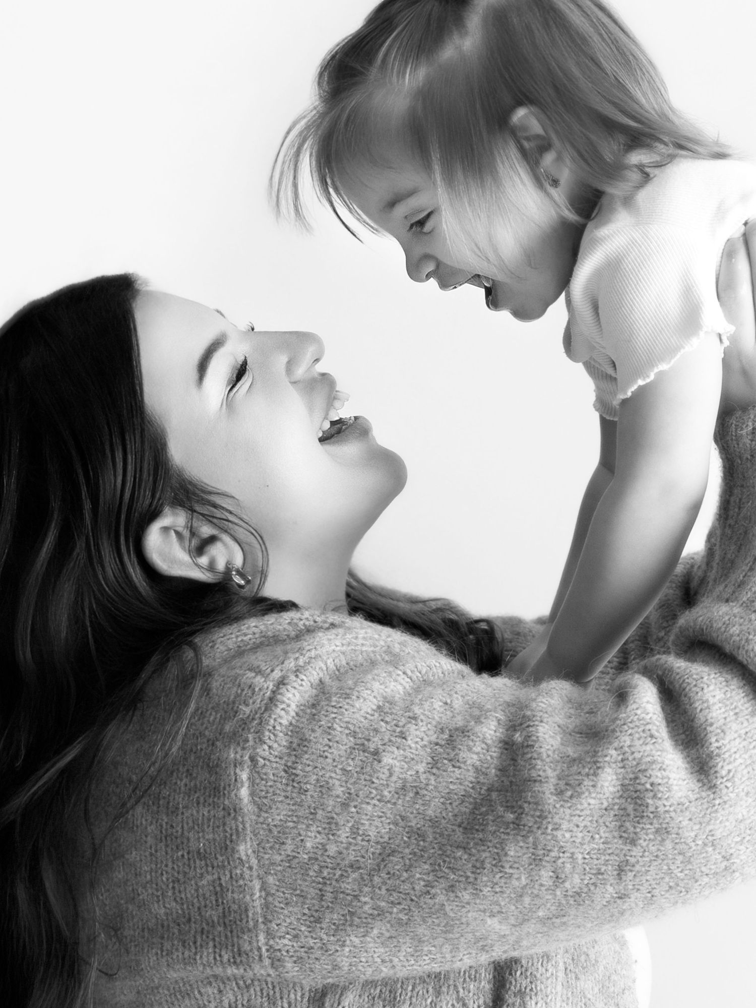 Black and white photo of a laughing mother lifting her joyful toddler daughter in a warm, tender moment.