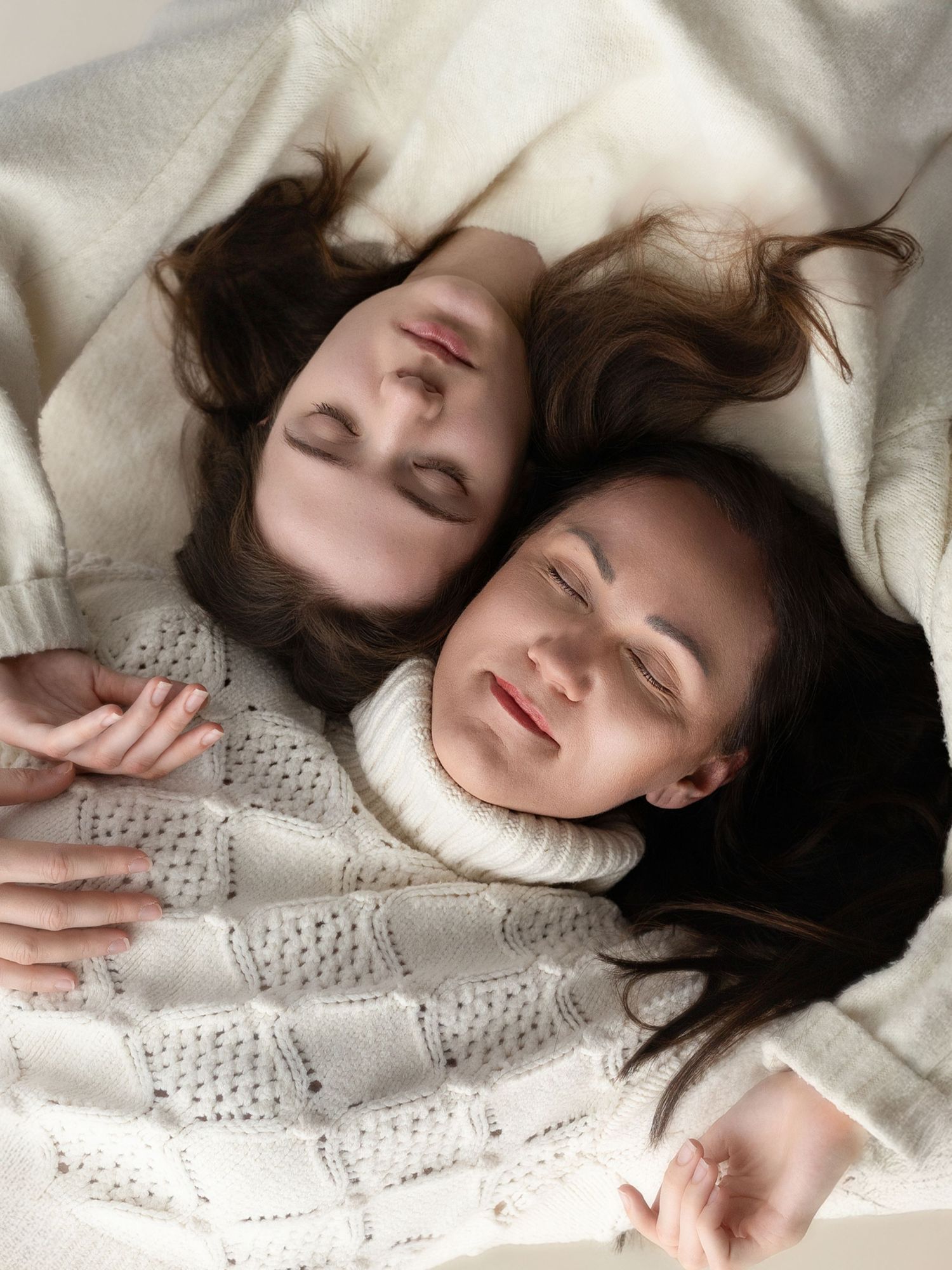 Two young women with closed eyes lying side by side on a white blanket, wearing cozy knit sweaters.