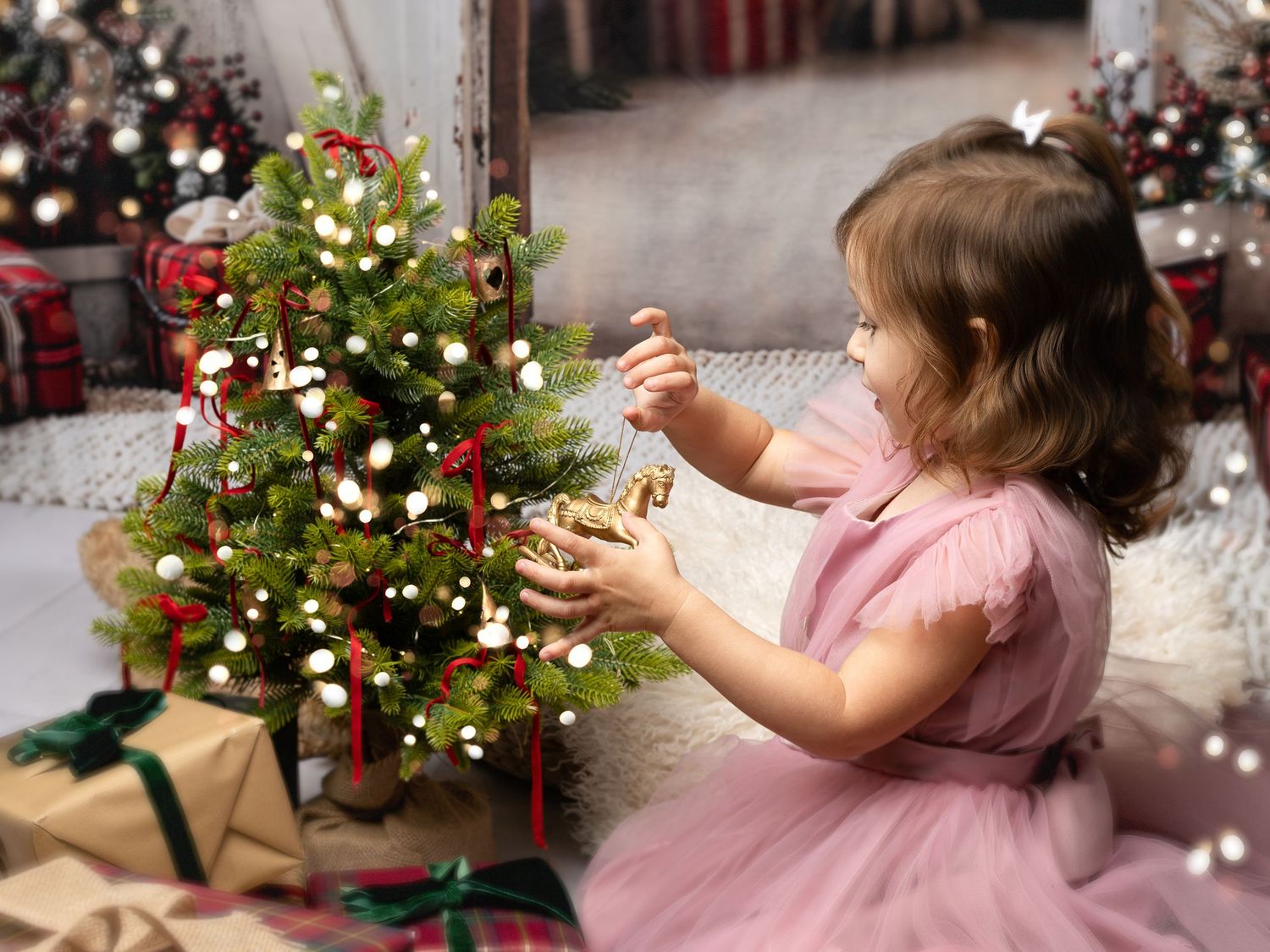 A young girl in a pink dress decorates a small Christmas tree surrounded by wrapped gifts and festive holiday decor.