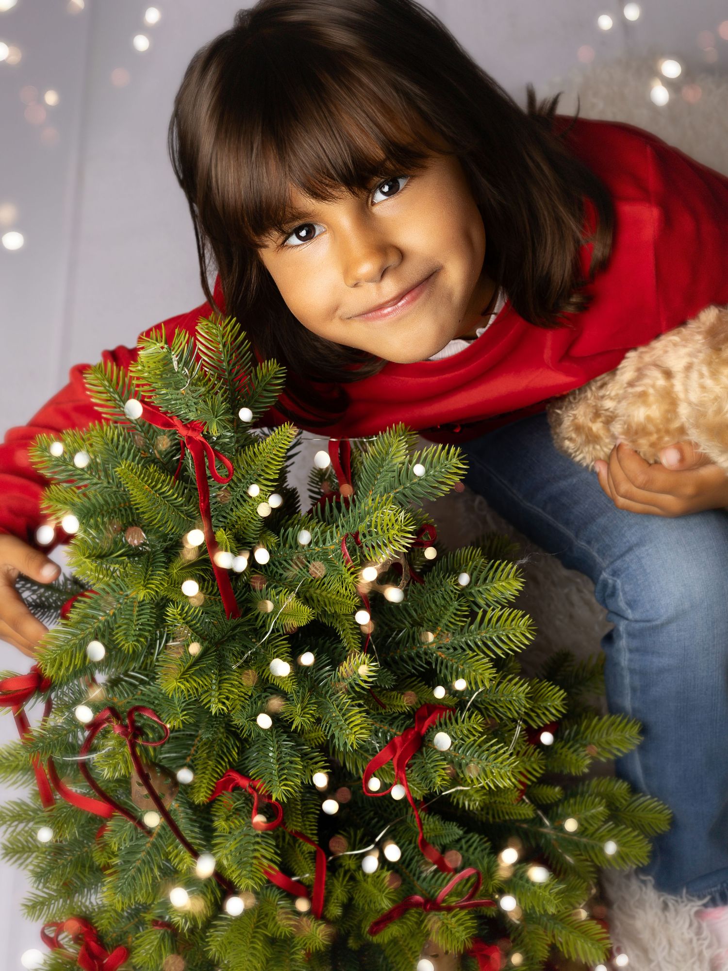A smiling young girl in a red sweater hugging a small decorated Christmas tree with white lights and red ribbon.