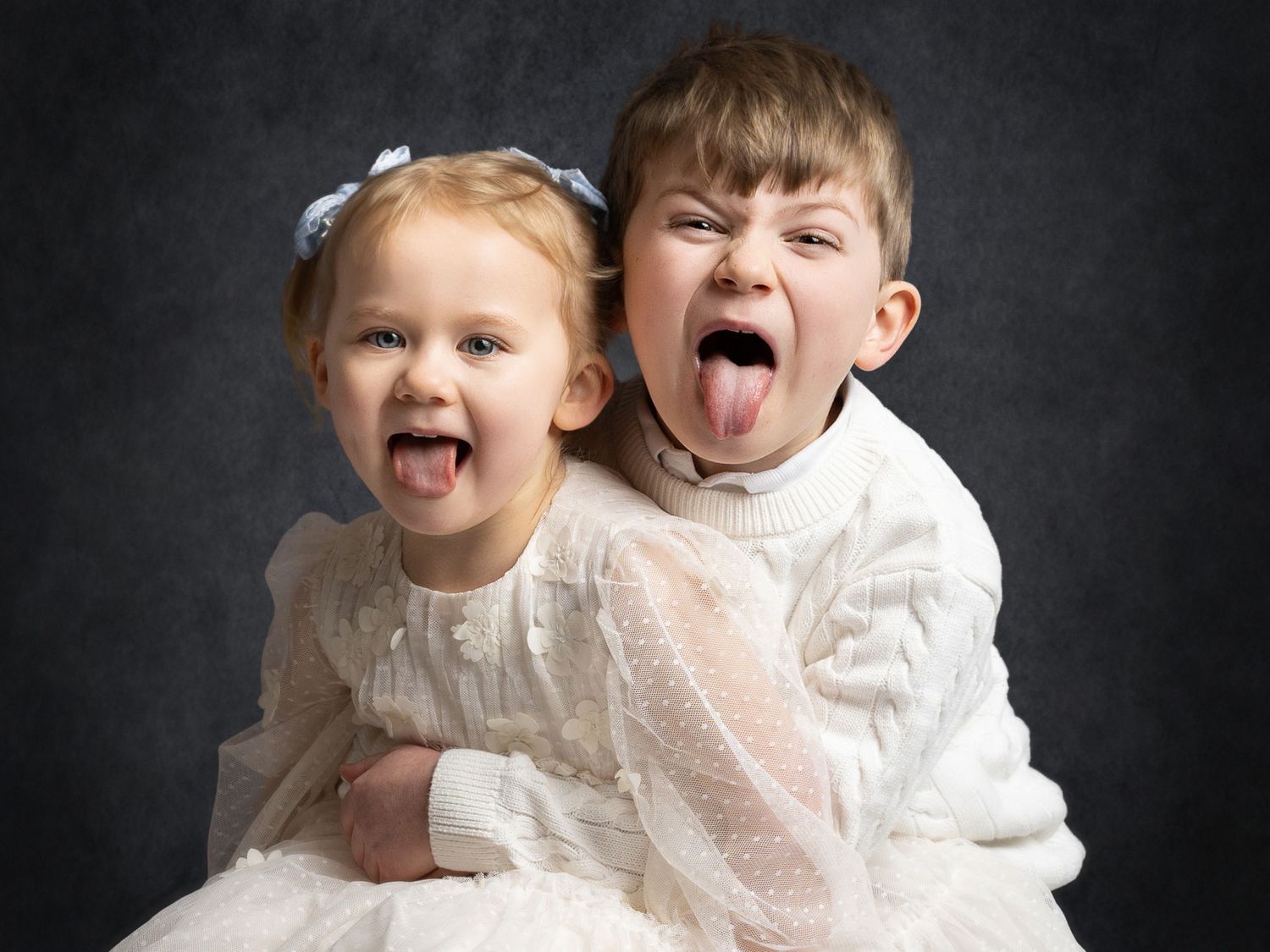 Two young children in white outfits playfully sticking out their tongues while posing together for a portrait photo.