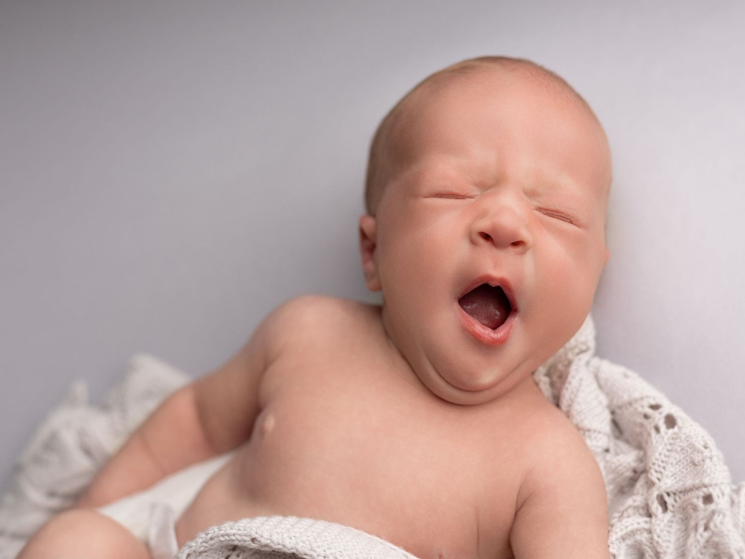 Adorable newborn baby yawning with eyes closed, wrapped in a white lace blanket on a gray background.