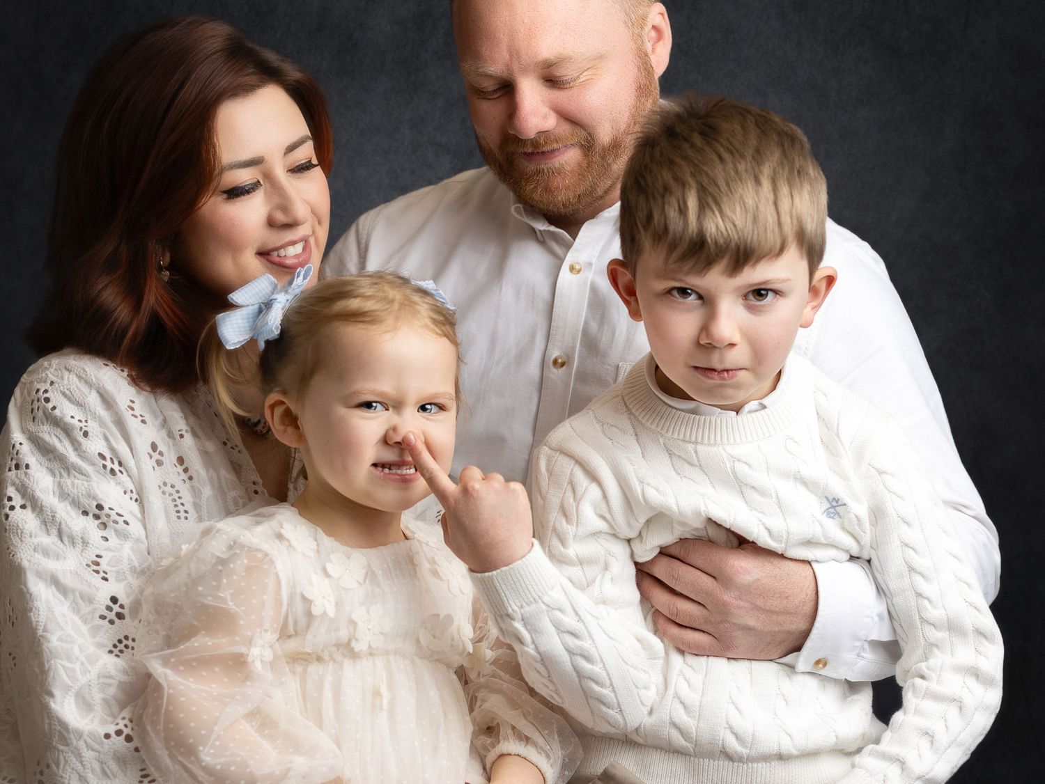 Family portrait of parents with two young children in white outfits against a dark studio background.