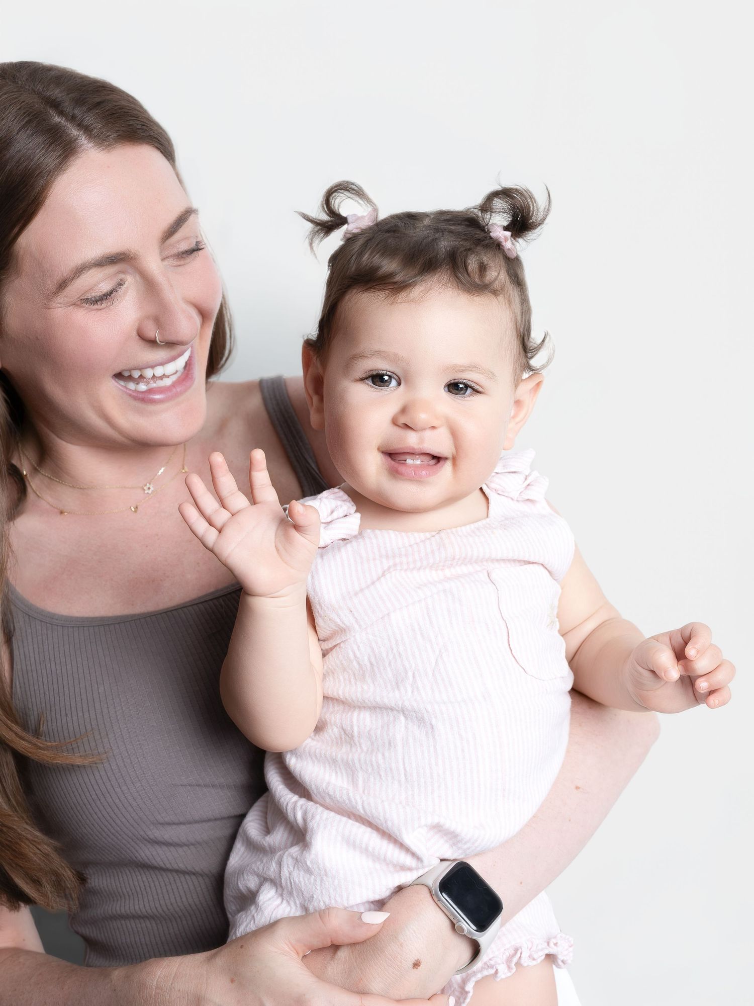 A smiling woman holds a happy baby girl with pigtails in a pink outfit, both posing against a white background.