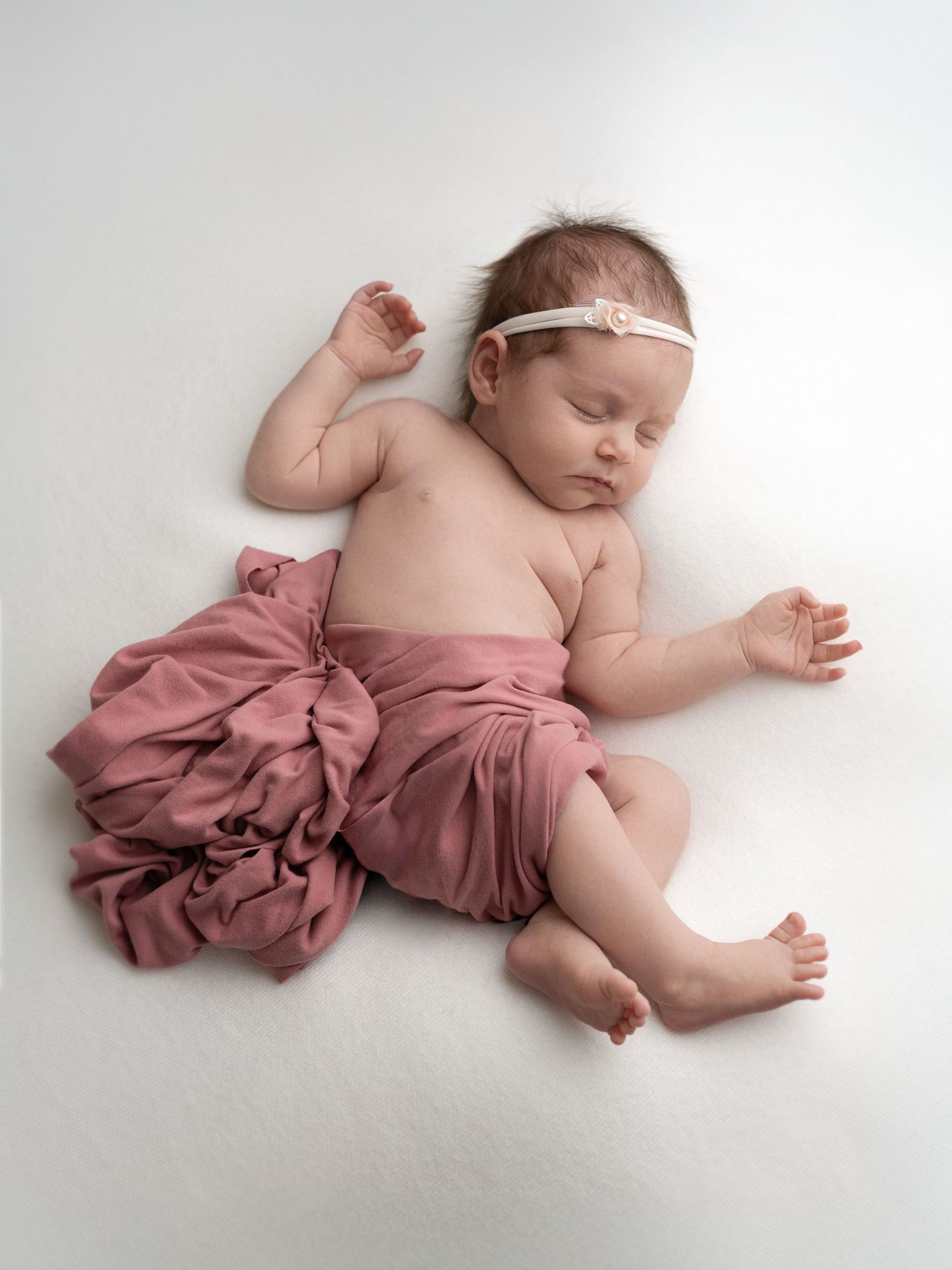 Newborn wearing dusty rose fabric lies peacefully on white background.