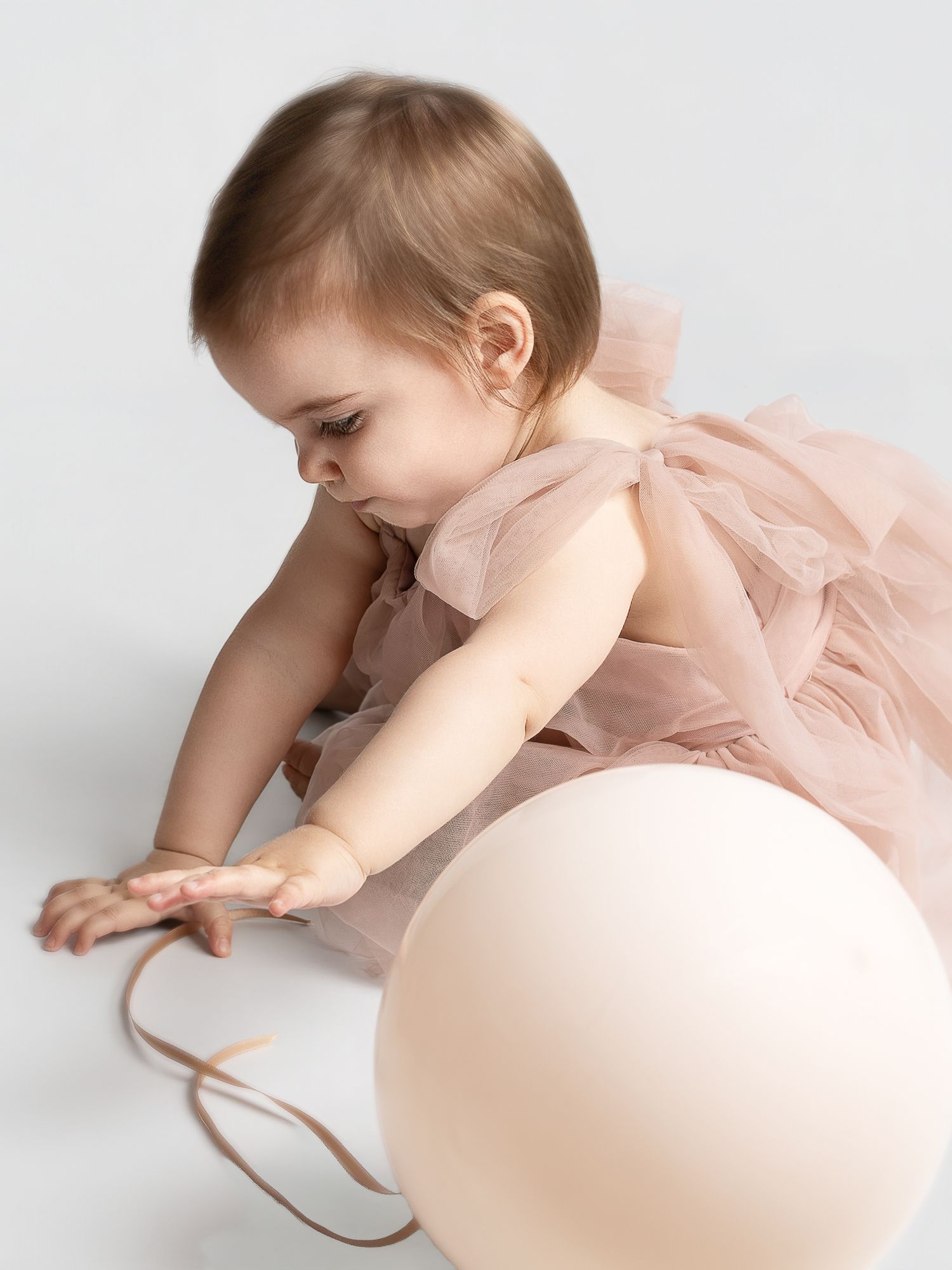 A baby girl in a pink tulle dress reaches for a cream balloon on a white background.