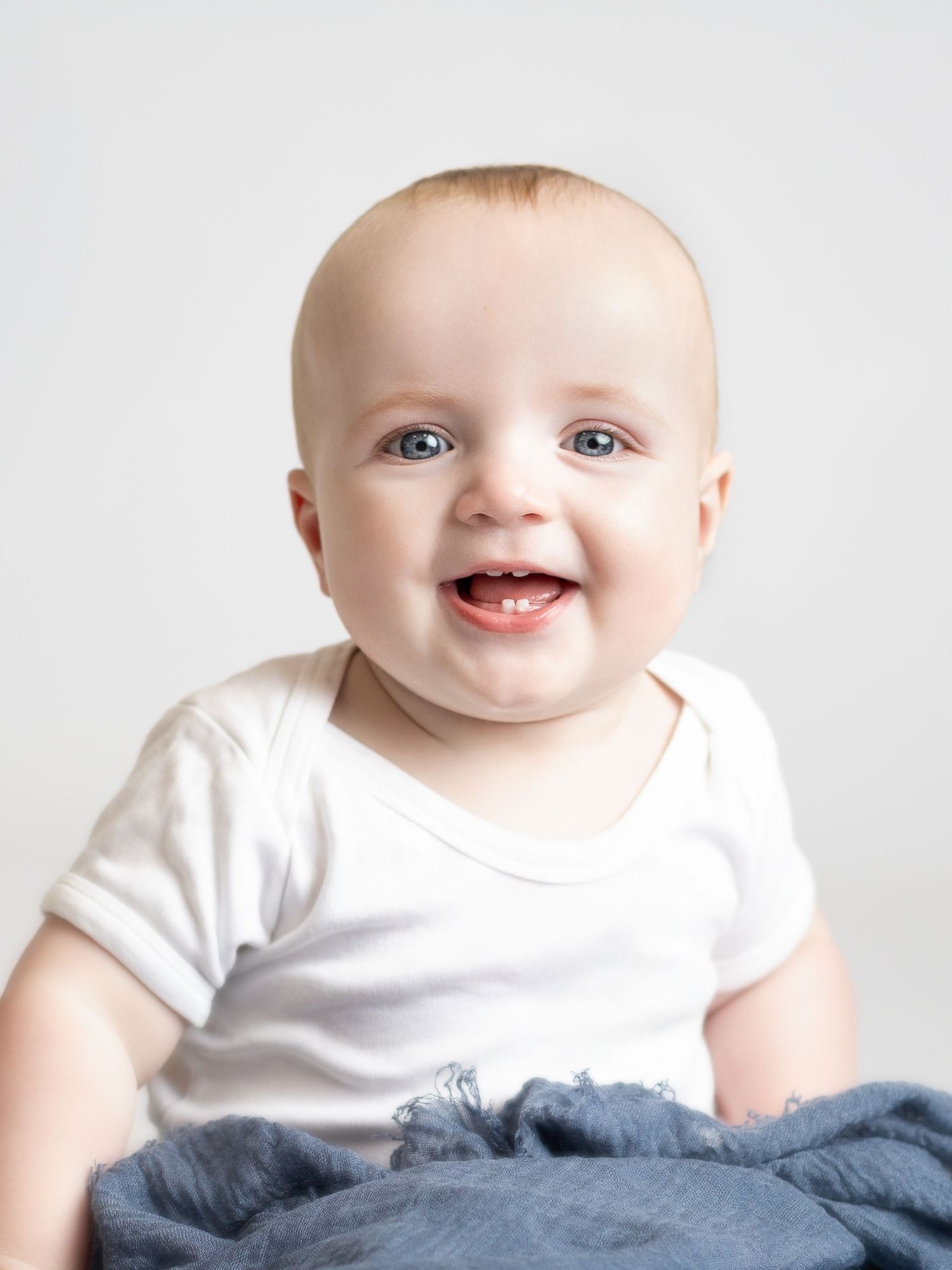 Smiling baby with blue eyes wearing a white onesie, sitting on a blue blanket against a light gray background.