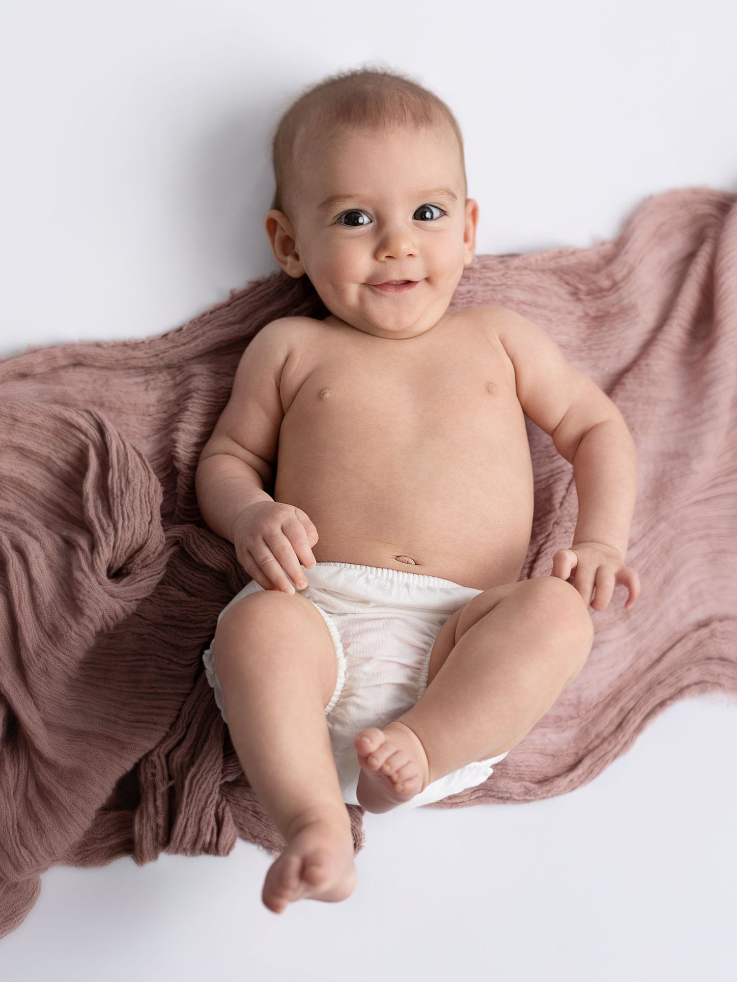 Smiling baby in a white diaper lying on a pink muslin blanket against a white background.
