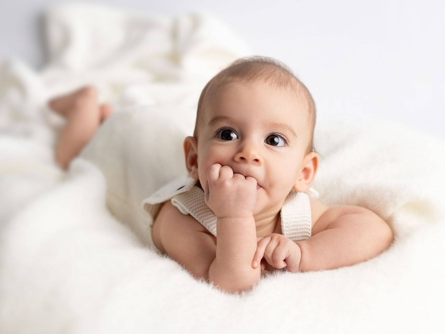 Adorable baby lying on white bedding with hand in mouth while looking up at camera.