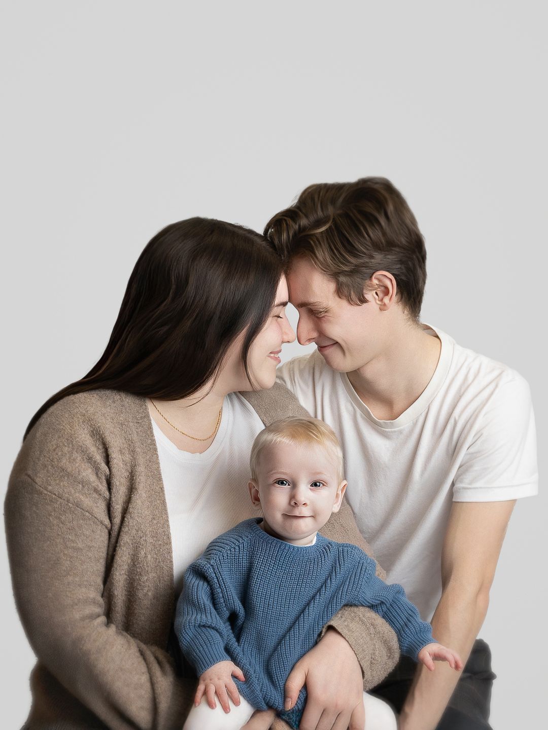 Young couple smiling at each other while holding their happy baby in a blue sweater against a light gray background.