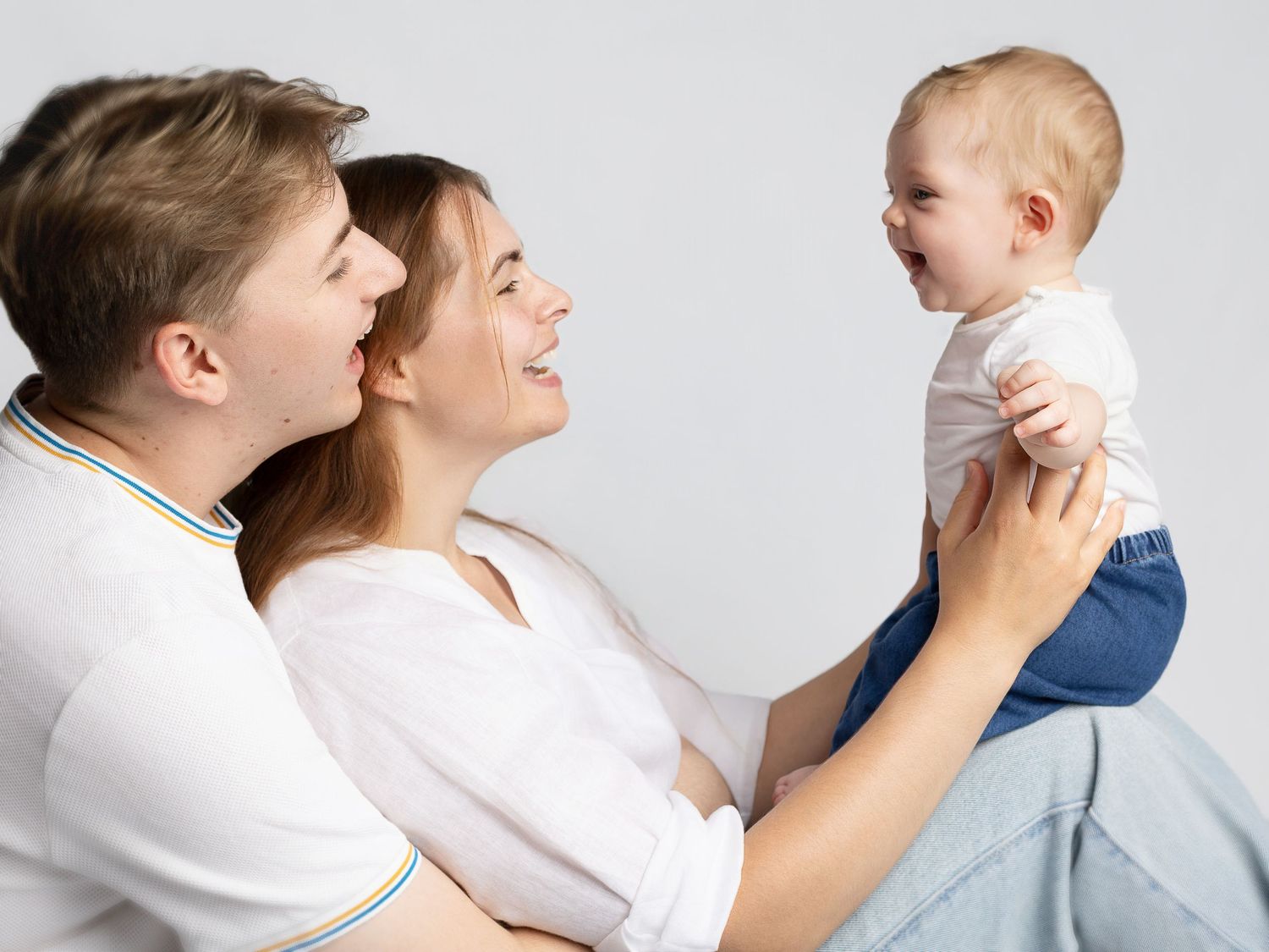 Happy young family with parents smiling at laughing baby in white studio setting.