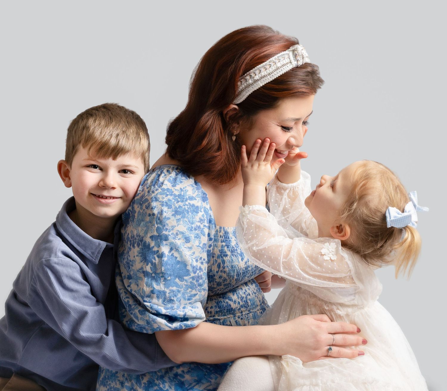 A mother in a blue floral dress smiles as her toddler daughter touches her face, while her young son hugs her side.