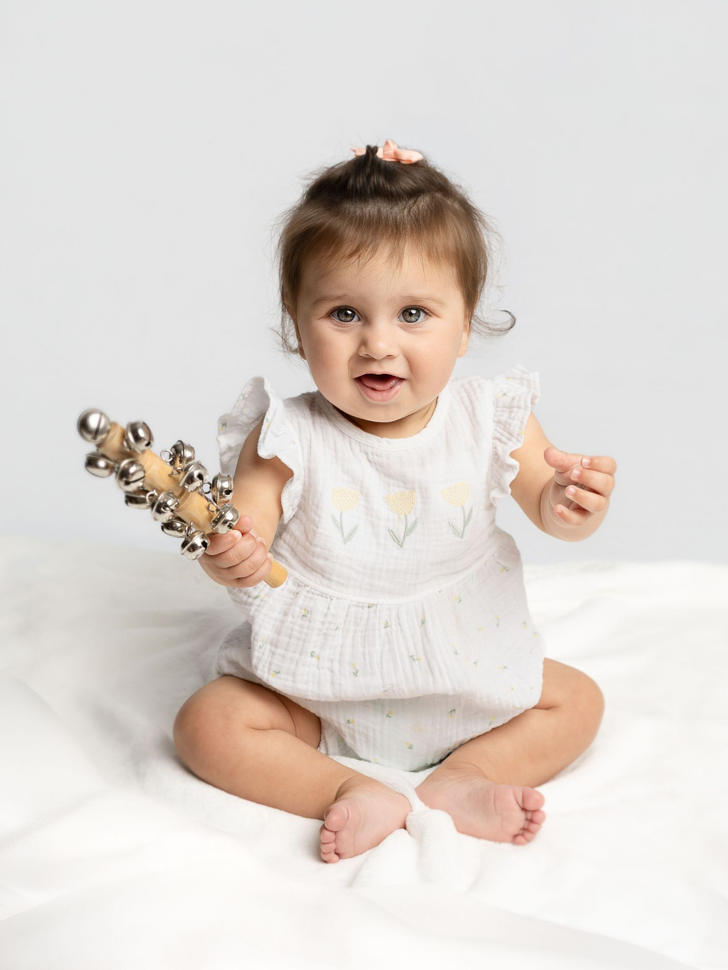 Smiling baby girl in white floral romper sitting on white blanket, holding a jingle bell stick toy.