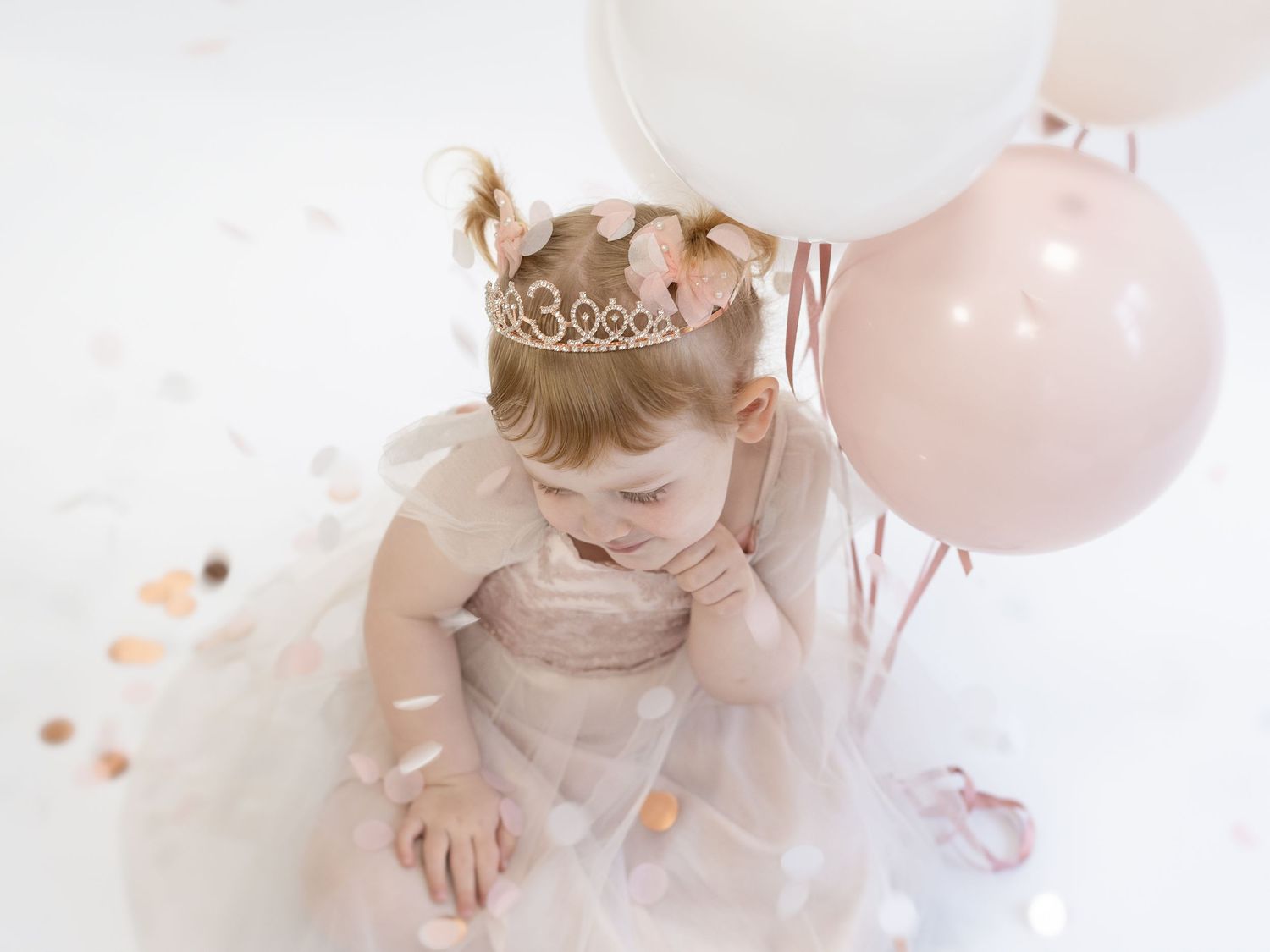 A small child in a sparkly dress and crown sits surrounded by pink balloons and delicate tulle decorations.