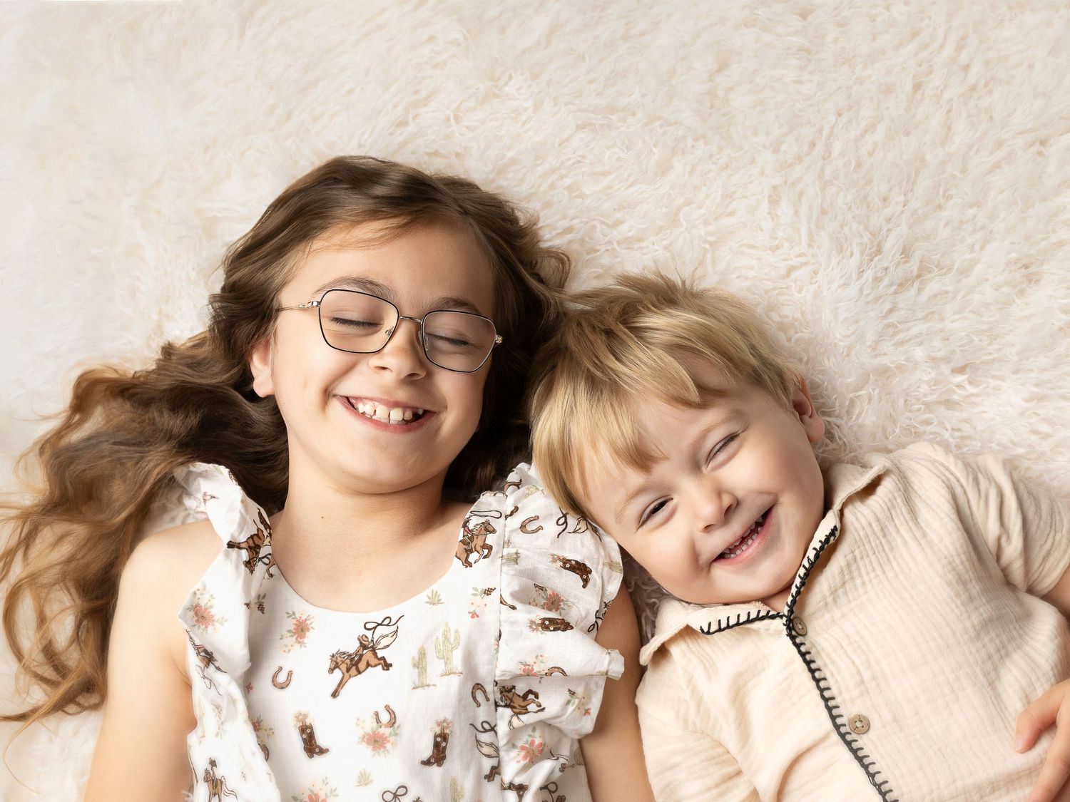 Two happy children lying on a fluffy white rug, smiling joyfully, girl with glasses and younger toddler sibling.