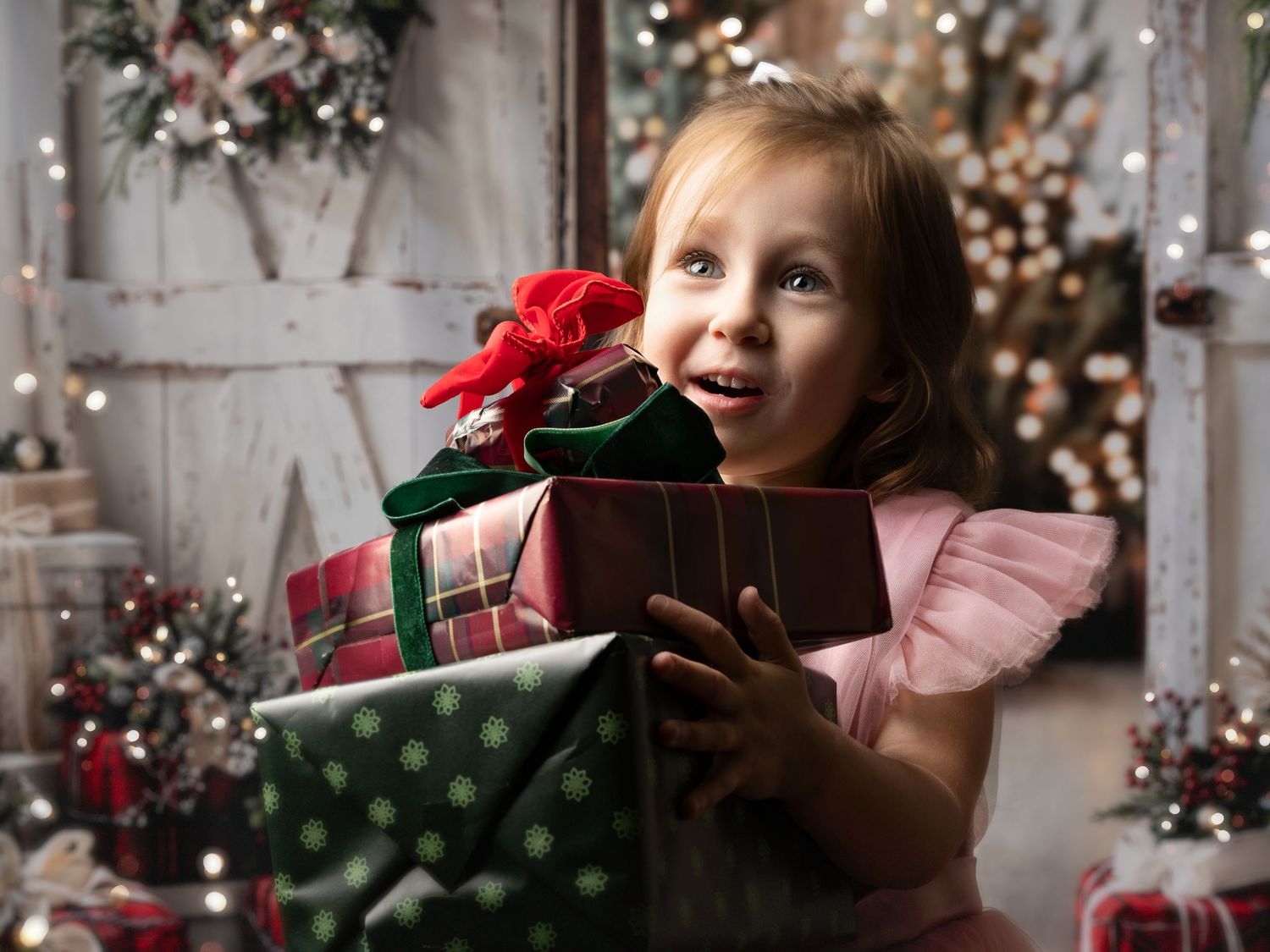 Excited little girl in pink dress holding Christmas gifts near decorated tree with twinkling lights.