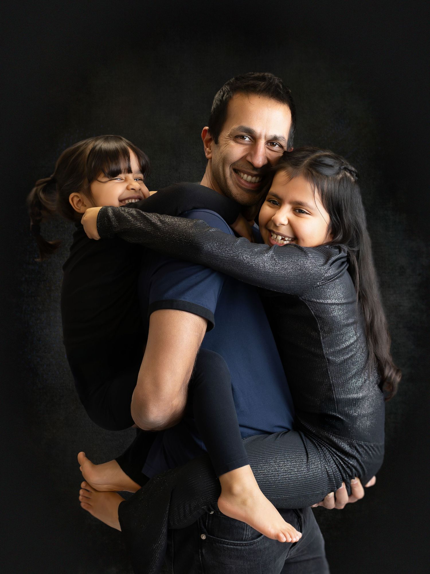 A smiling man holds two young girls in his arms against a dark studio background.