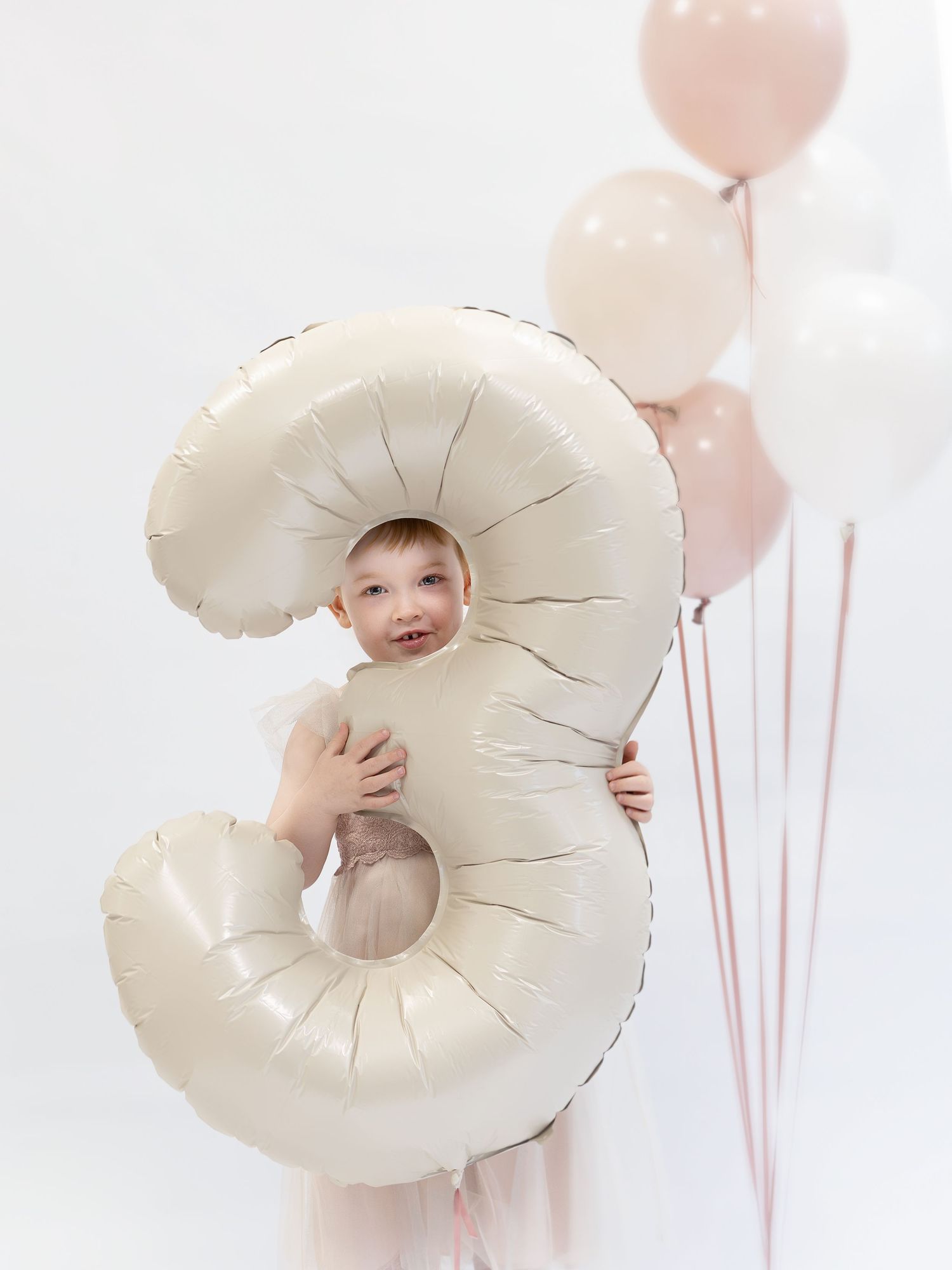 Young child holding a large cream number 3 foil balloon with pink balloons on white background.