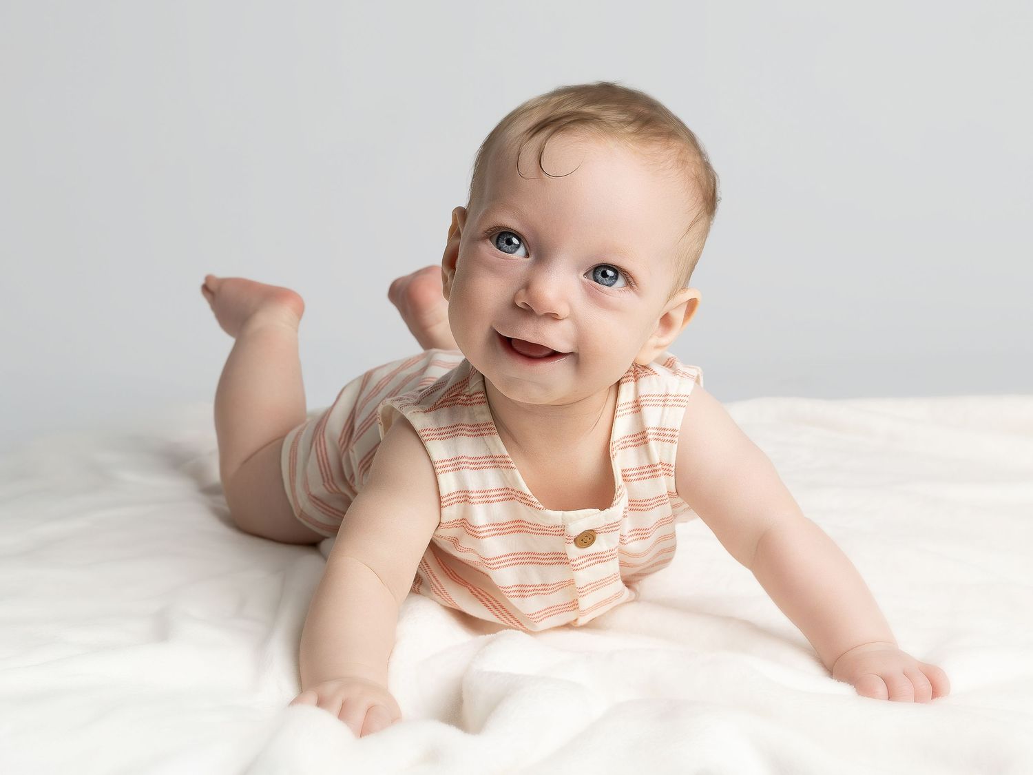 Smiling baby in striped onesie doing tummy time on white blanket against gray background.