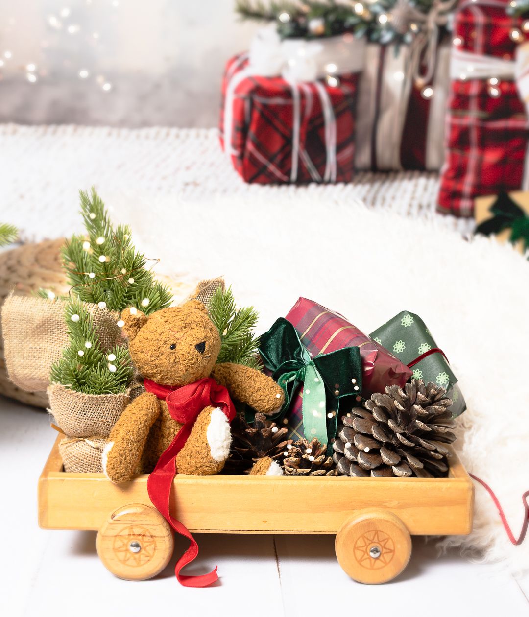 A wooden toy wagon filled with a teddy bear, pine cones, ribbons, and mini Christmas trees in a festive holiday setting.