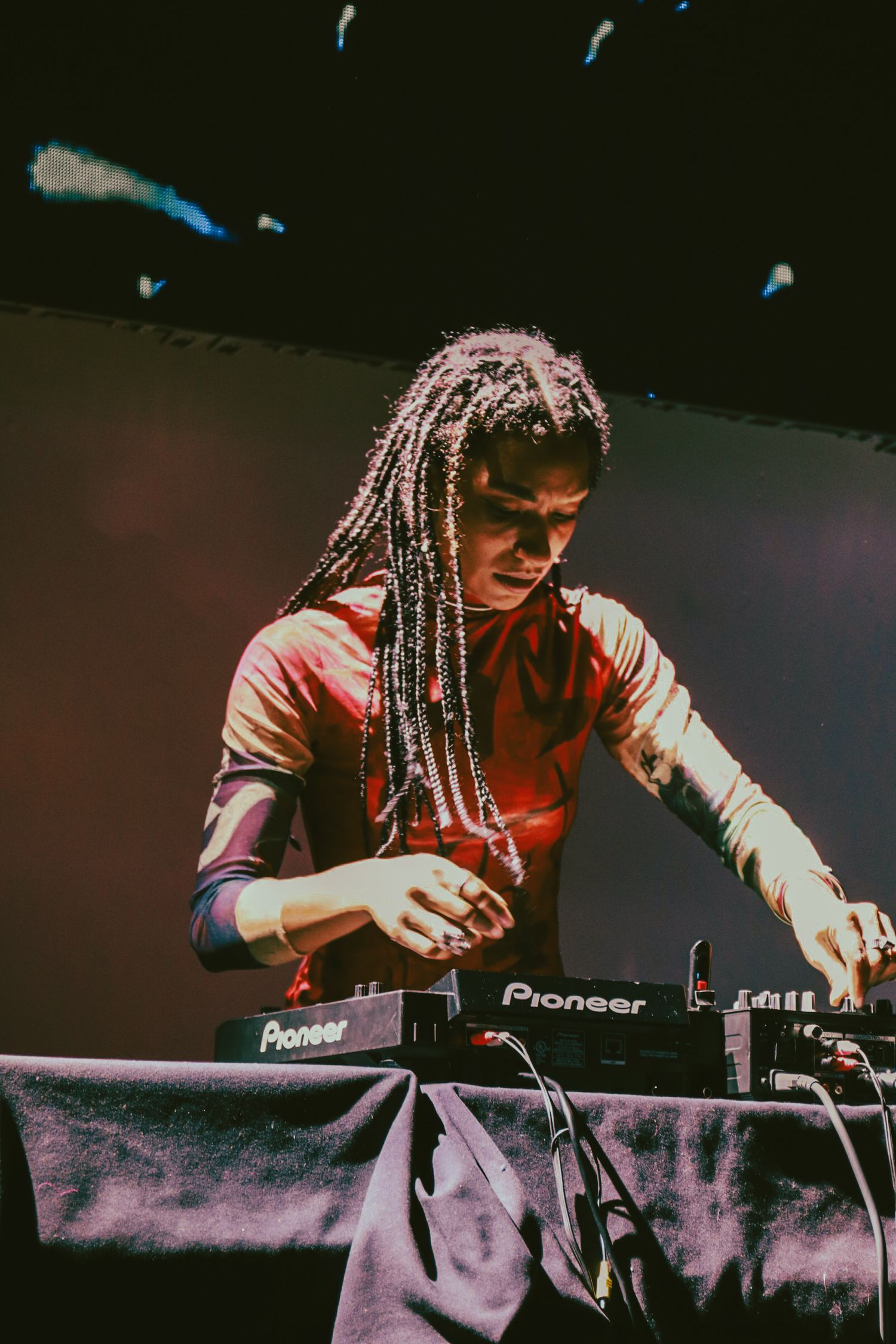 DJ with long braids performing on Pioneer decks under dramatic stage lighting at a live music event.
