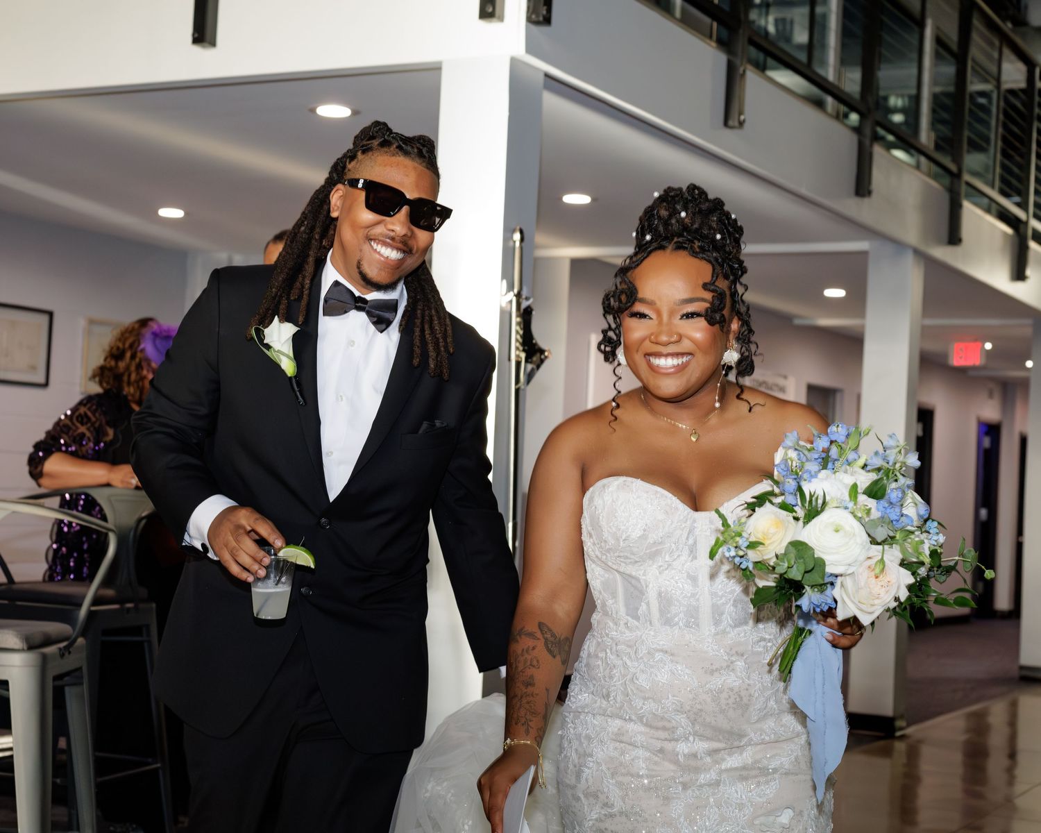 Bride in strapless gown holding blue bouquet walks with groom in black tuxedo and sunglasses at wedding.