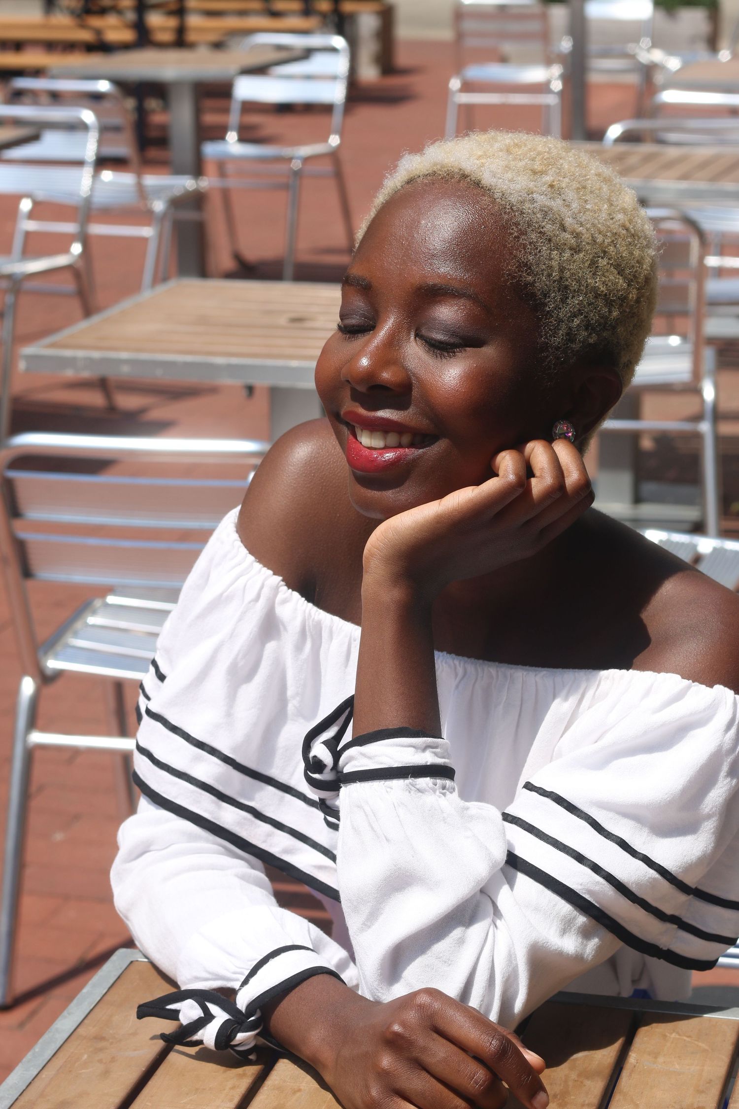 Smiling woman with short blonde hair in white off-shoulder top sits at outdoor table in sunlight.