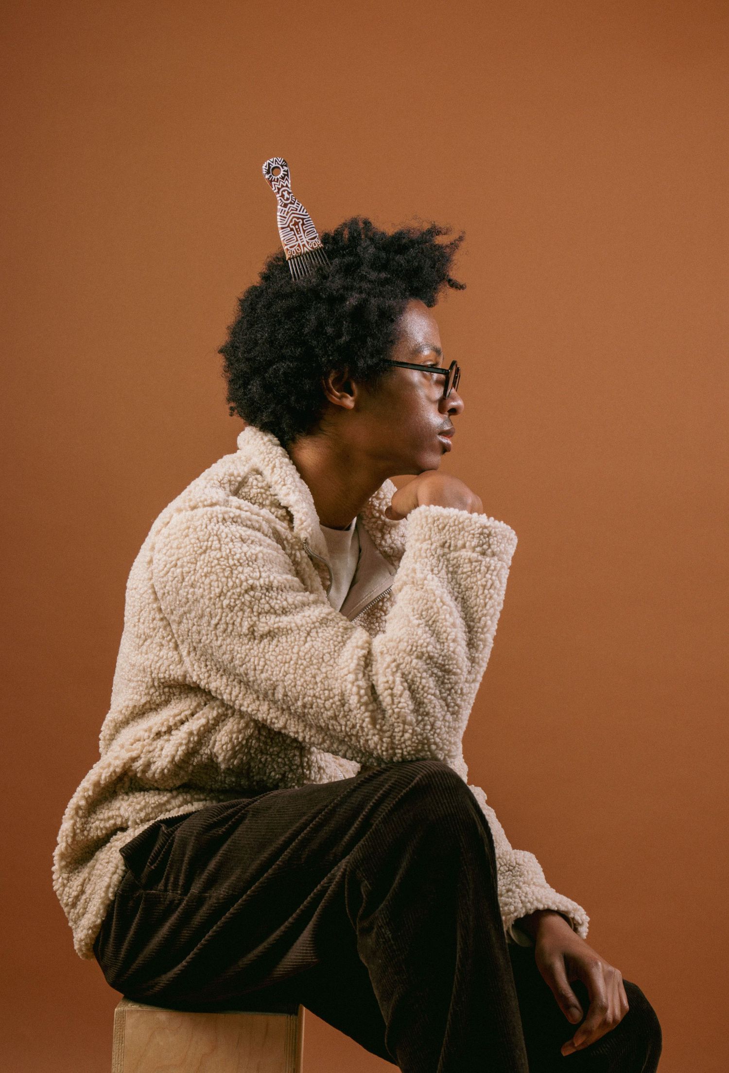 Young Black man with glasses and natural hair sits thoughtfully wearing cream fleece jacket on brown background.