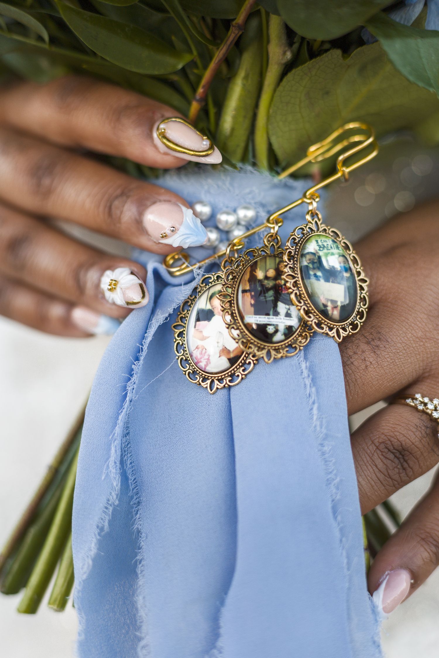 Close-up of bridal bouquet with gold oval photo charms and blue ribbon held by manicured hands.