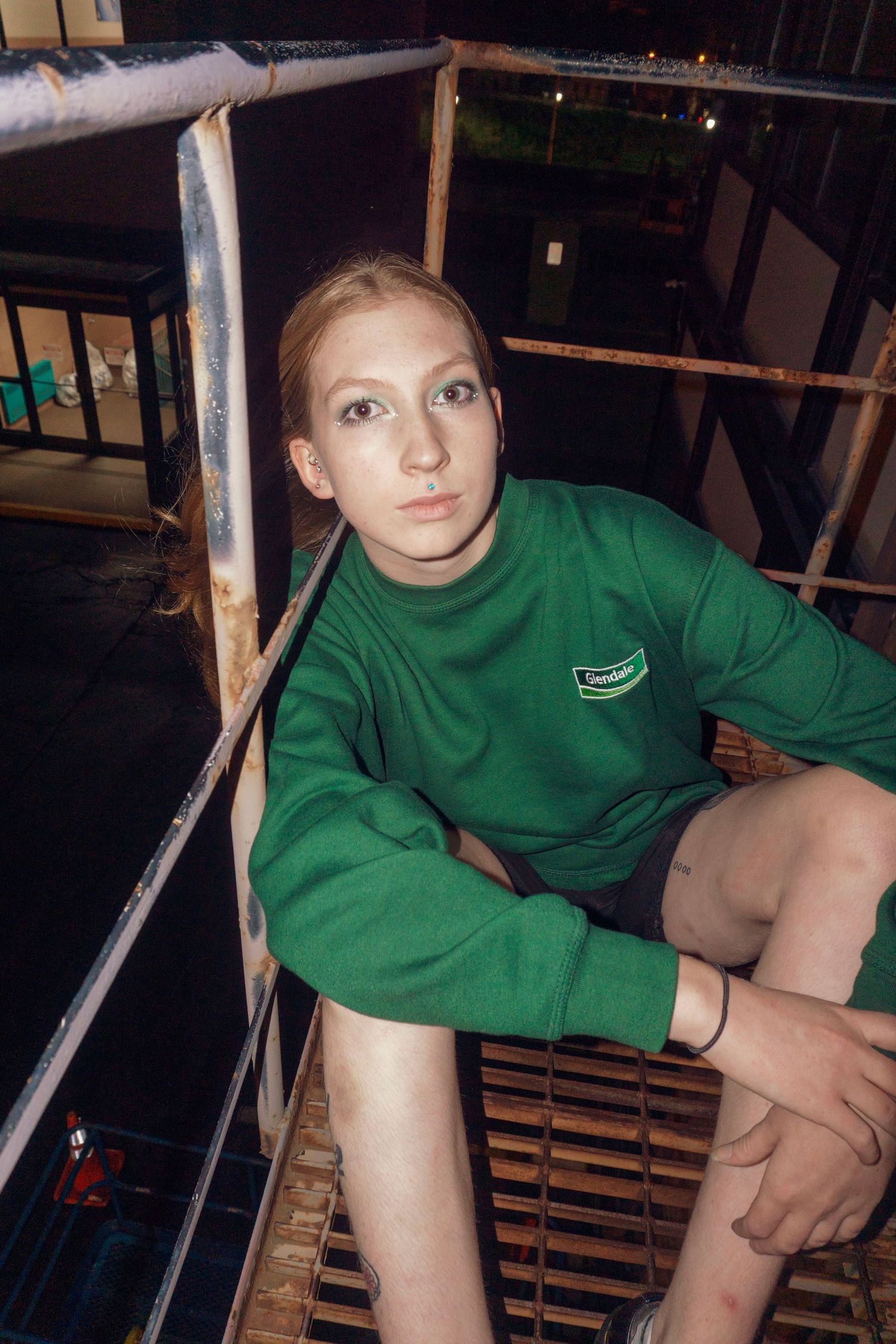 Young woman in green Lacoste sweatshirt sits on metal grating at night looking up.