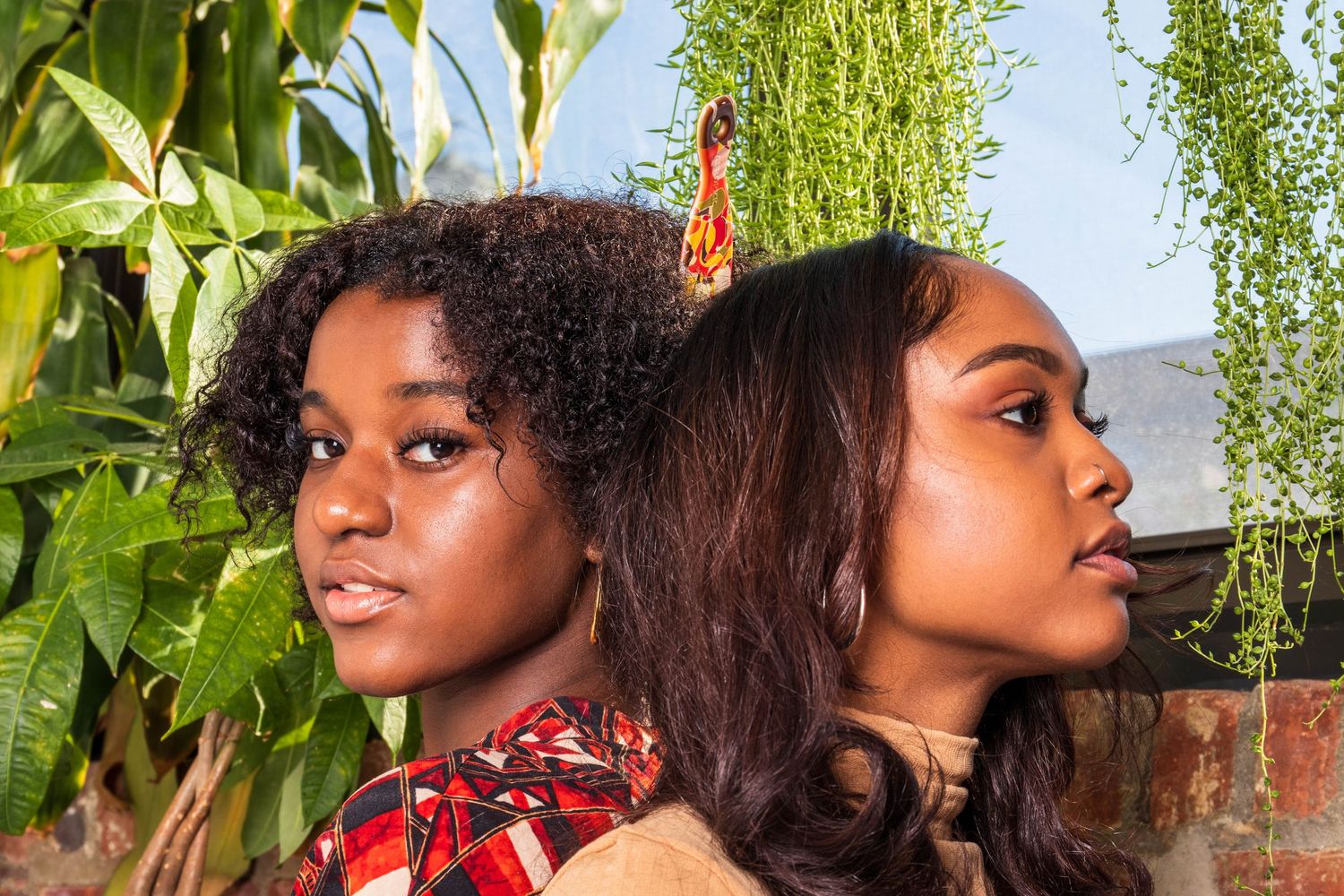 Two young Black women pose back-to-back in profile view surrounded by lush green tropical plants outdoors.