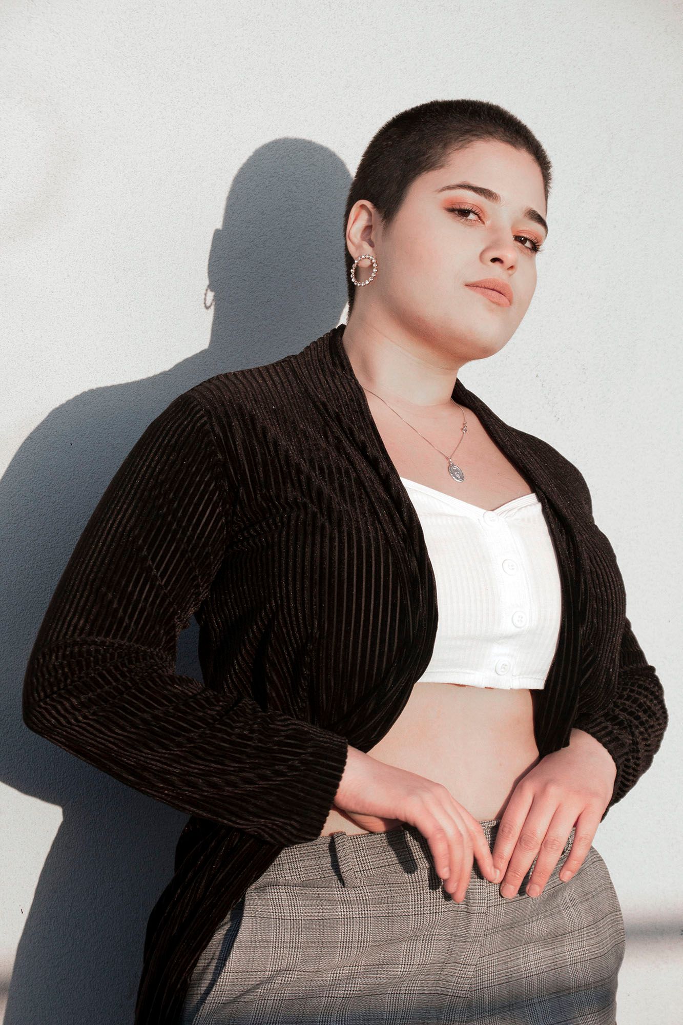 A woman with a shaved head wearing a black blazer and white crop top poses against a white wall.