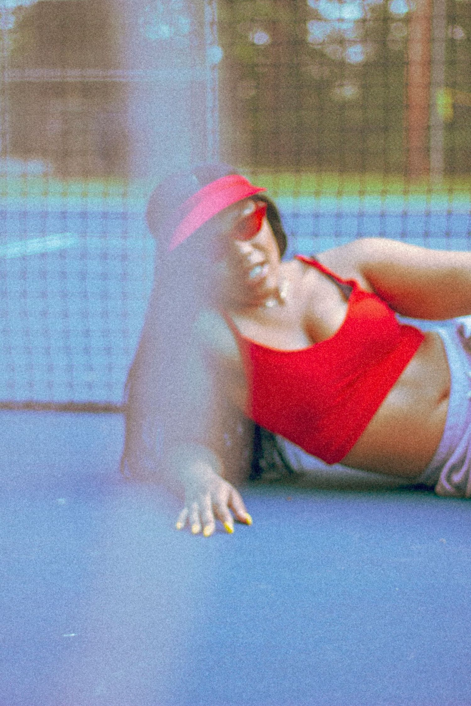 Woman in red crop top and pink visor lying on blue tennis court in film photo style.