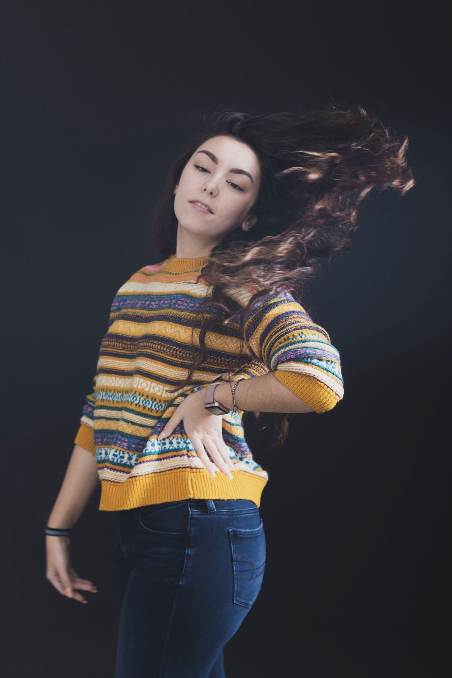 Young woman in colorful striped sweater and jeans with hair flowing dynamically against dark background.