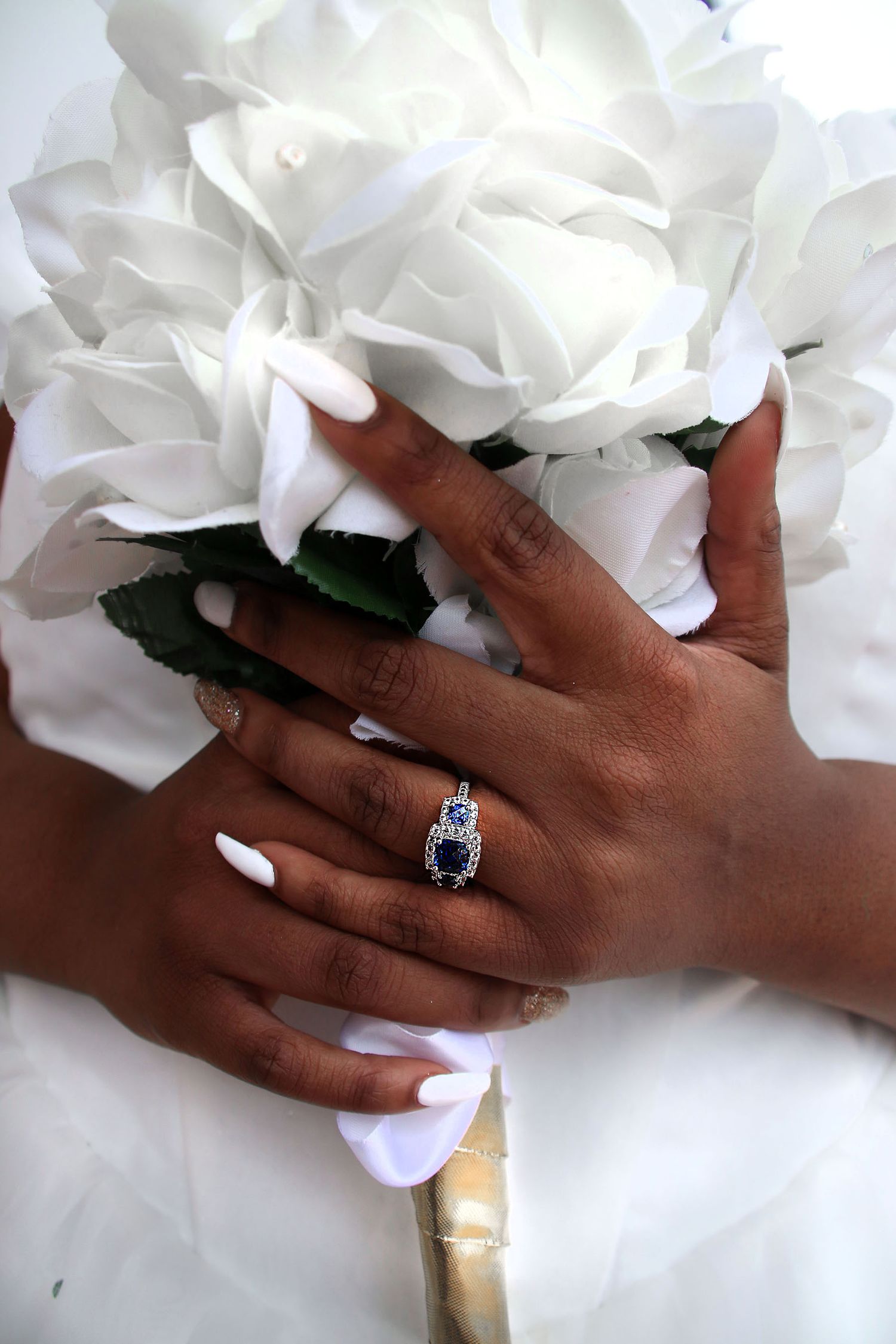 Bride holding white rose bouquet displaying sapphire and diamond engagement ring with white manicure.