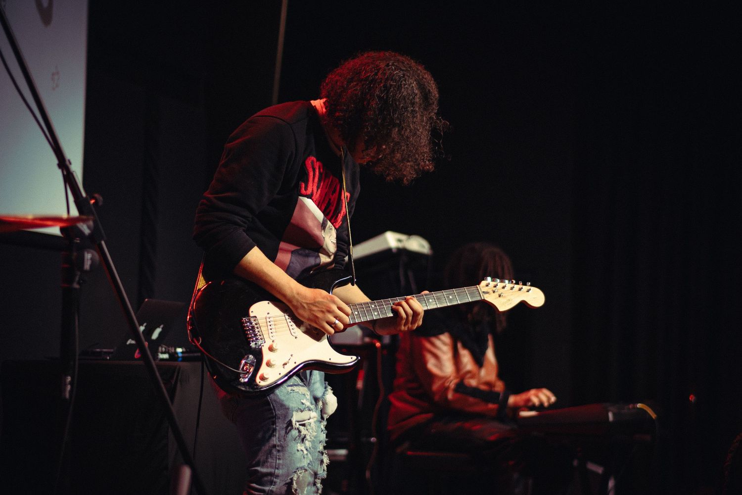 A curly-haired guitarist plays a white Fender Stratocaster on a dark concert stage.