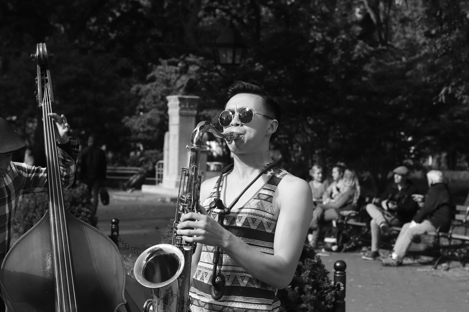 Black and white photo of a woman playing saxophone outdoors in a park wearing sunglasses and a patterned dress.