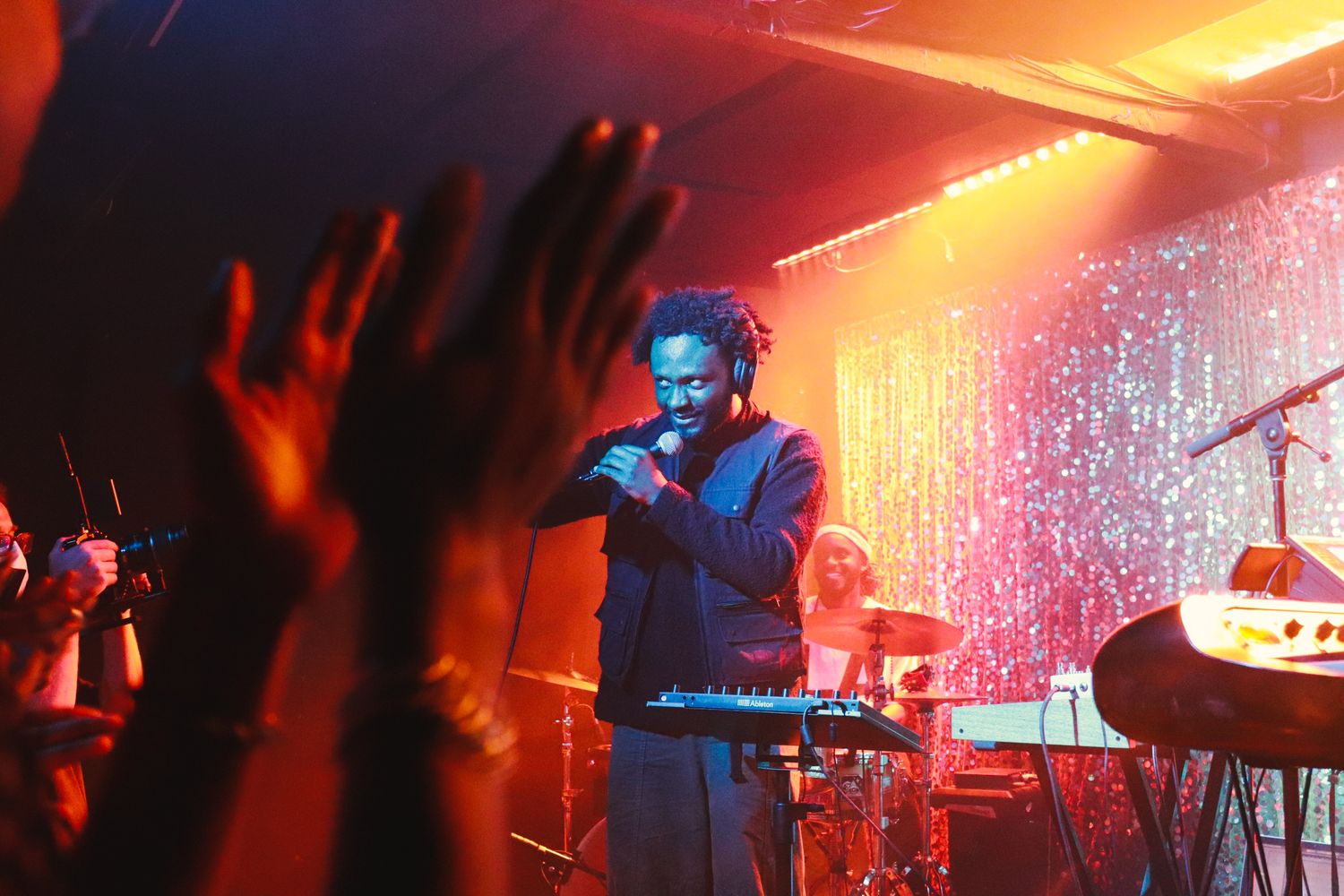 Male artist performing on stage with keyboard while audience raises hands under warm orange lighting.