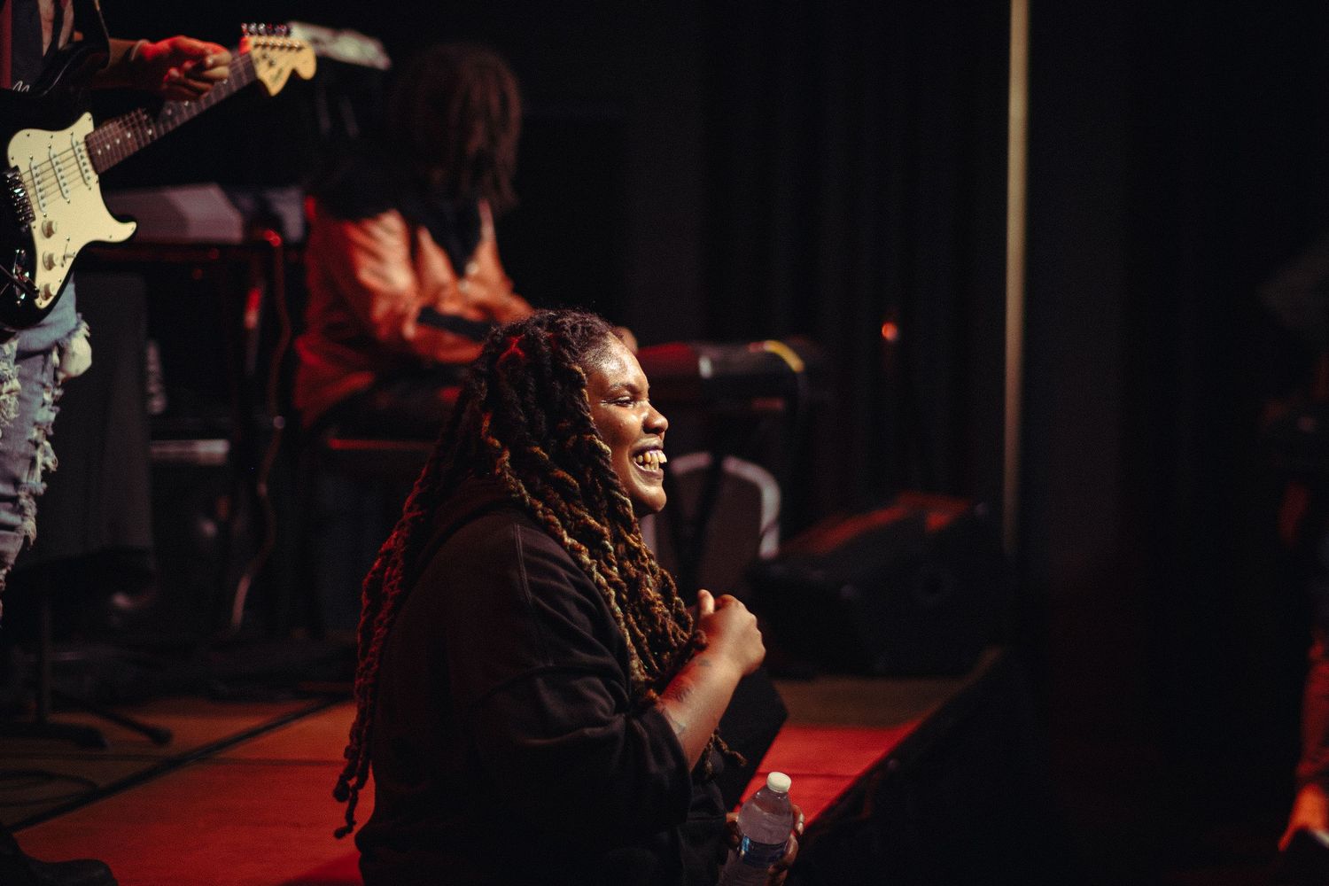 Woman with long locs smiling joyfully while sitting on a stage during a live performance.