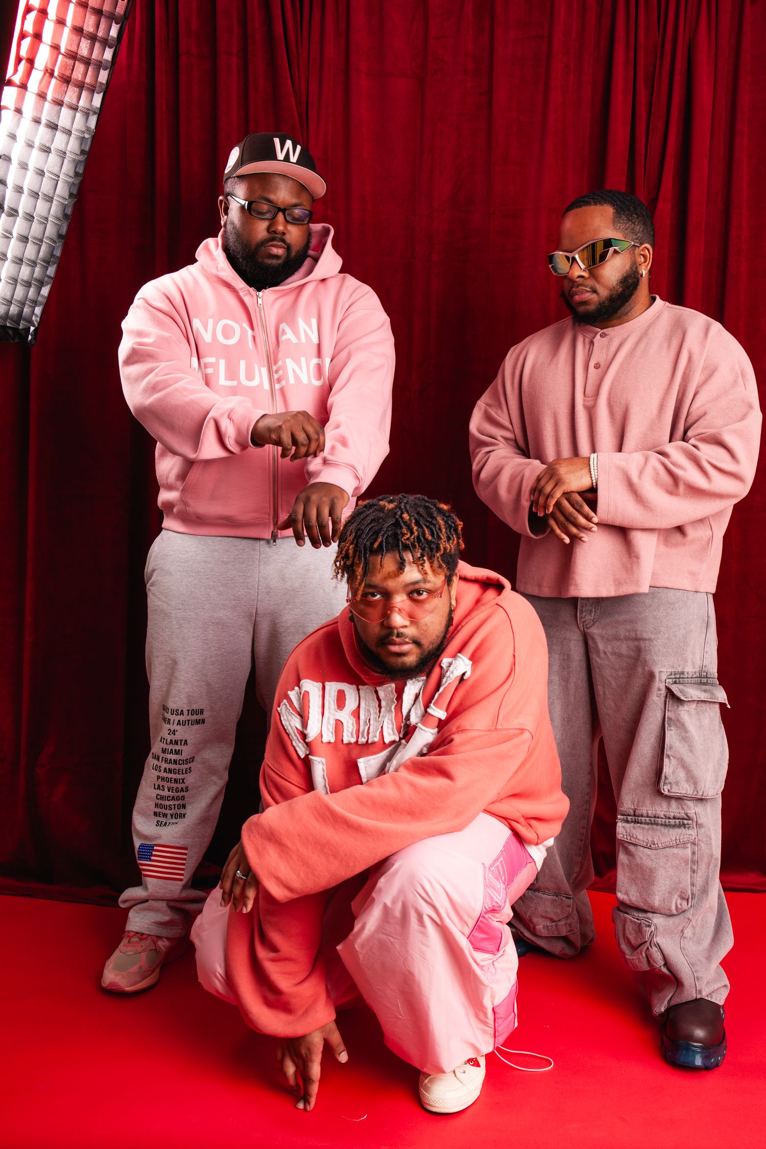 Three Black men in pink and grey streetwear pose together against red curtain backdrop.