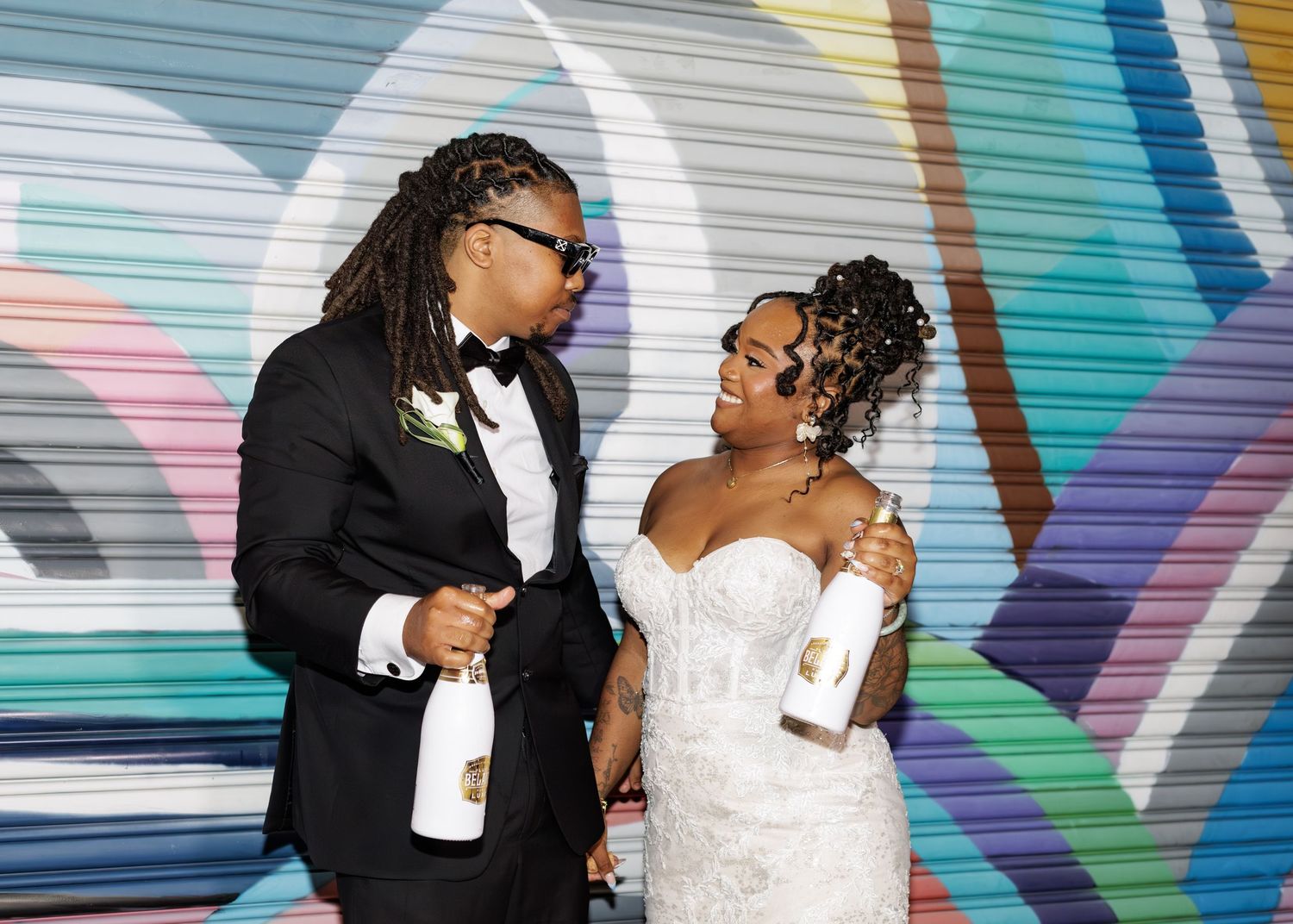 Newlywed couple laughing together holding champagne bottles in front of colorful graffiti wall.