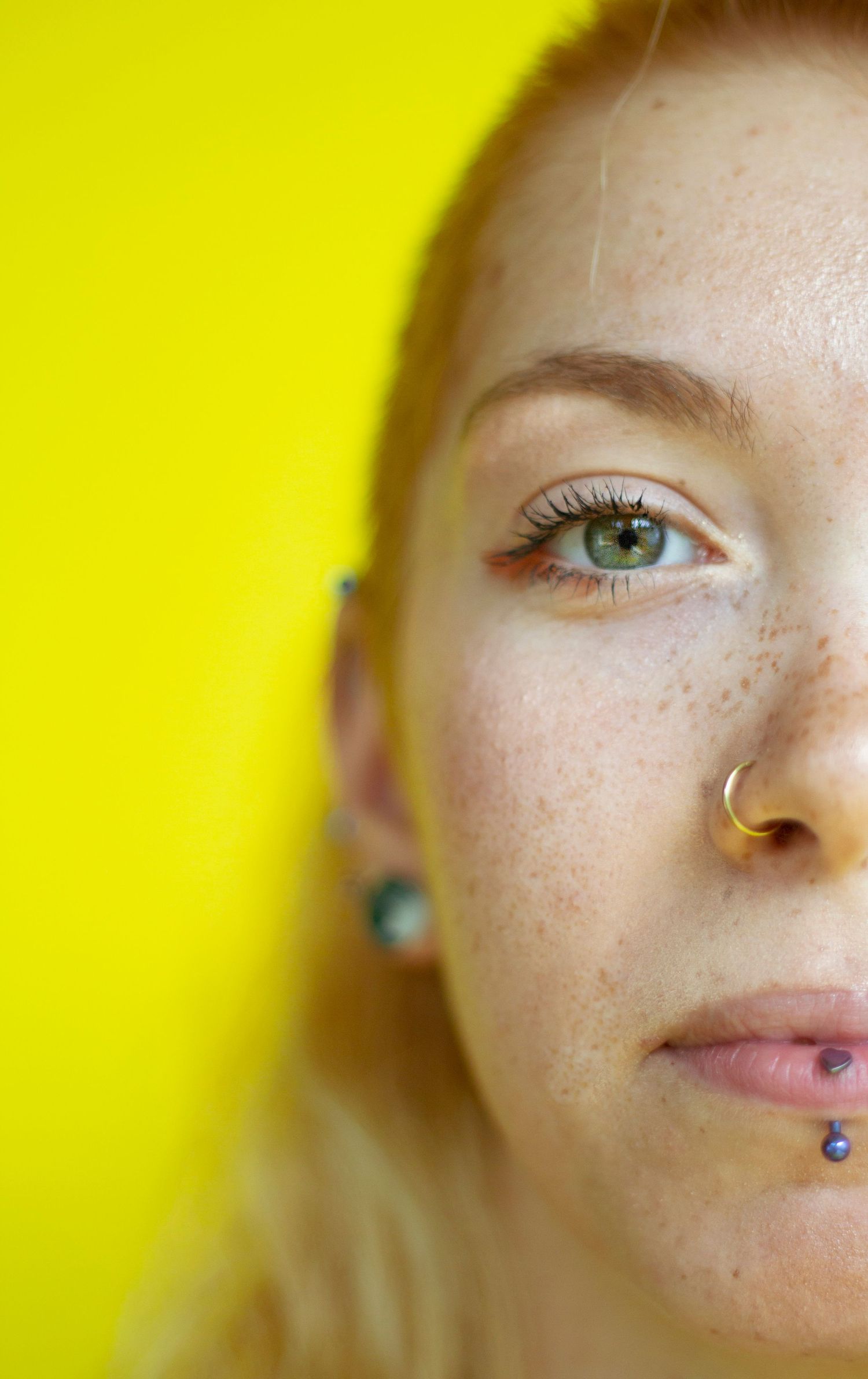 Close-up half portrait of freckled woman with nose and lip piercings on yellow background.