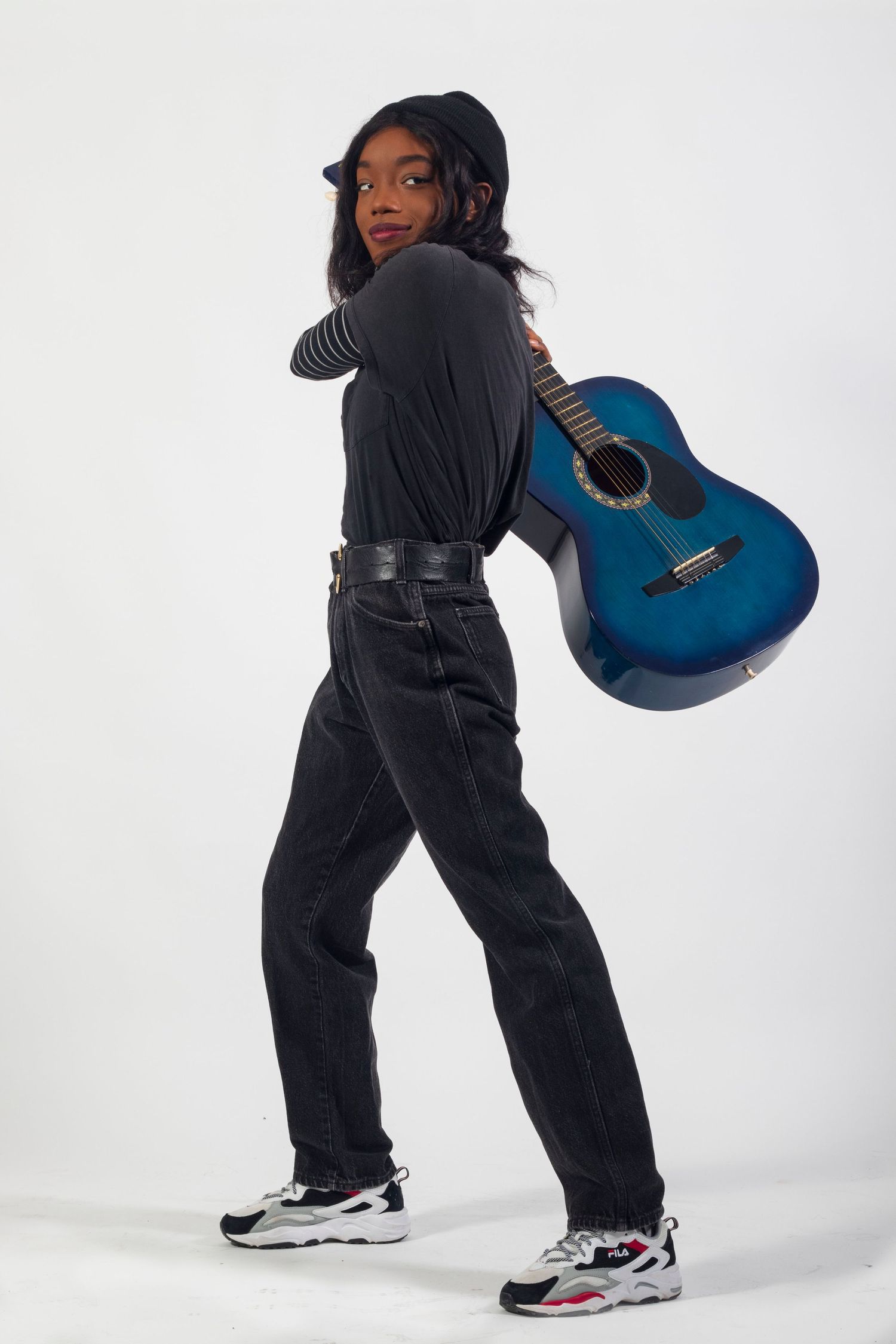 Young woman in all-black outfit holding blue acoustic guitar over shoulder on white background.