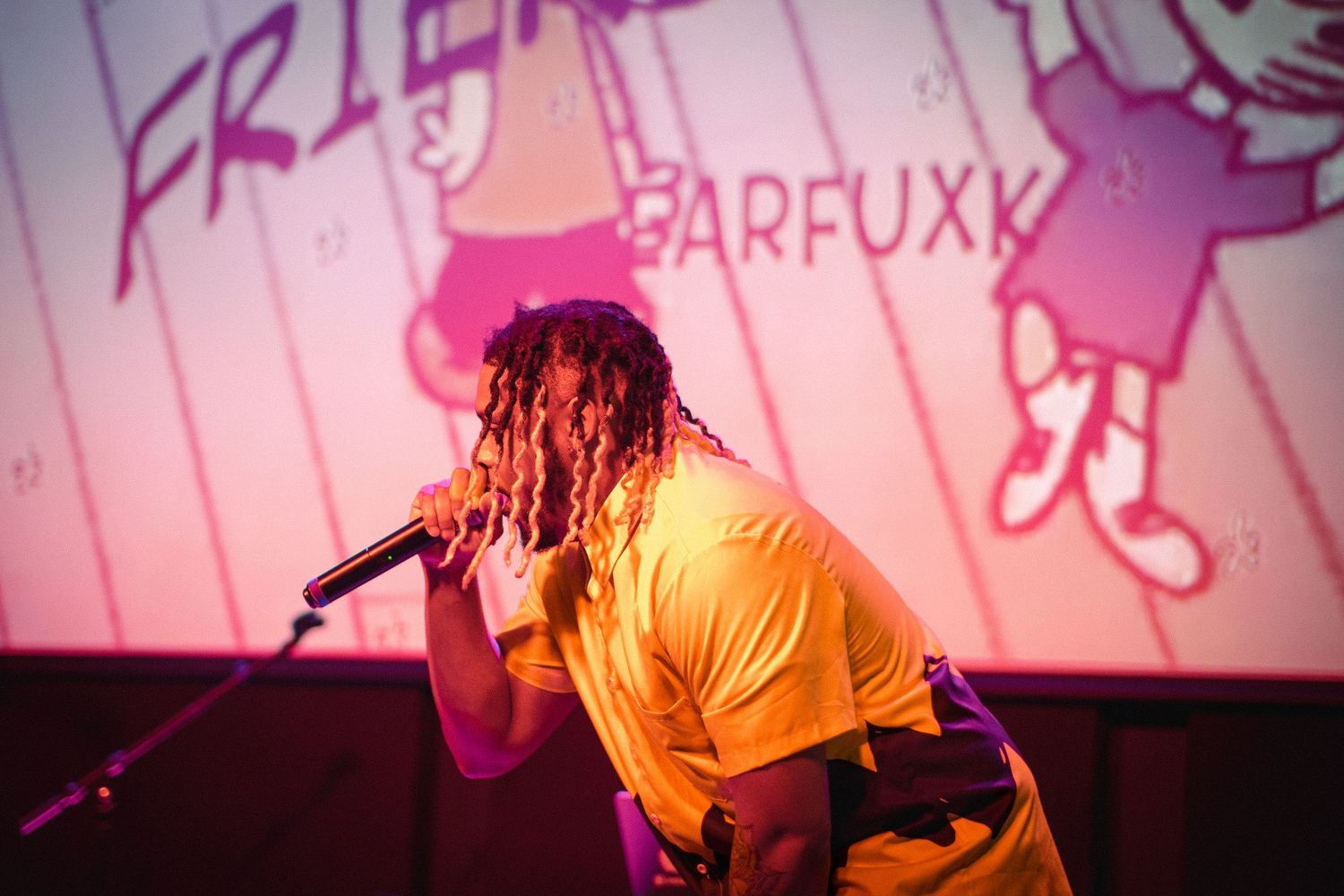 A performer in a yellow shirt raps on stage with colorful animated graphics projected behind him.