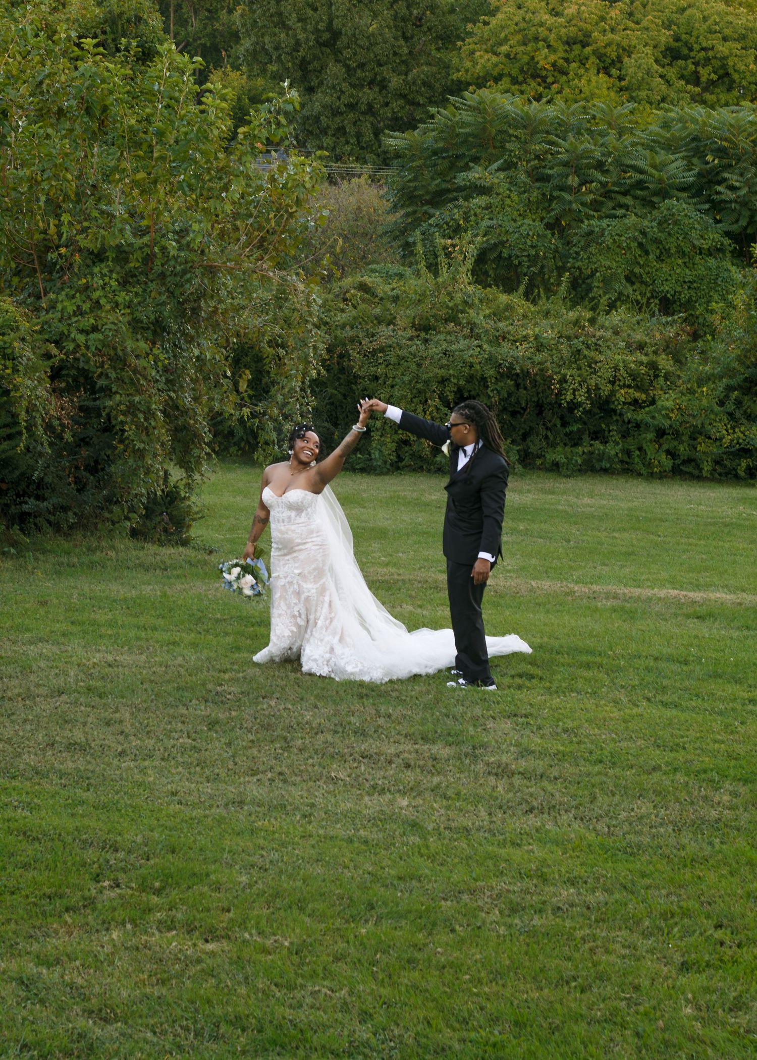 Bride in a white lace gown dances with groom in a black tuxedo on a green lawn outdoors.
